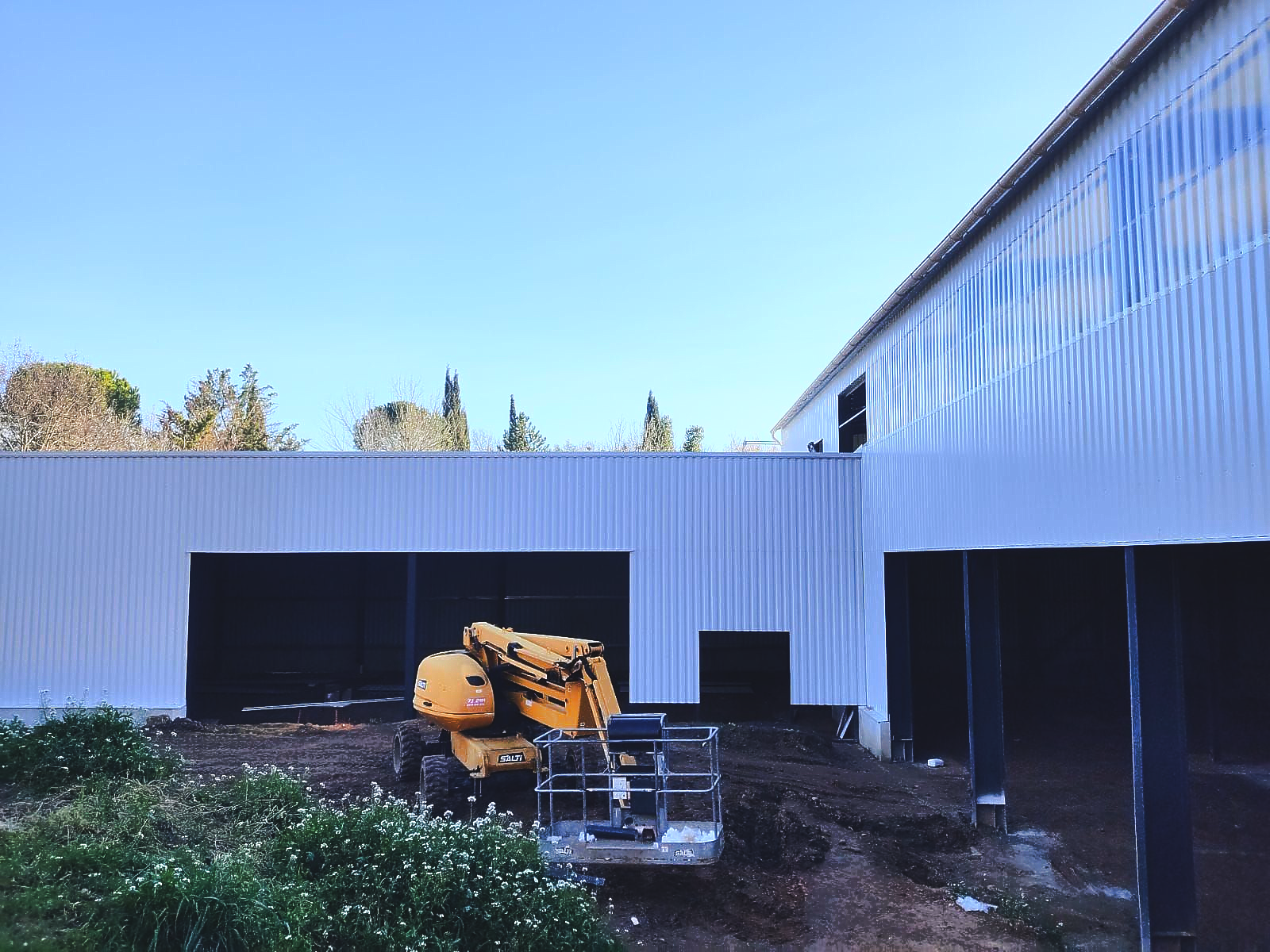Des engins de chantier jaunes dans une cour à côté d'un bâtiment métallique blanc sous un ciel bleu clair