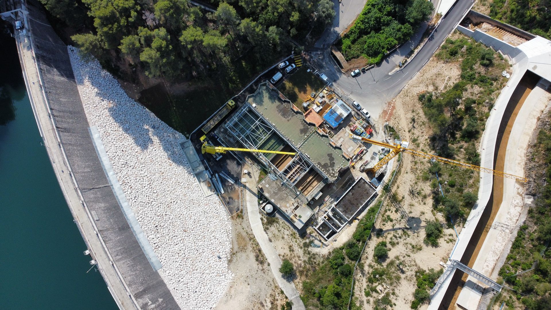 Vue aérienne d'un chantier de construction côtier, à proximité d'une digue et de l'eau, avec des arbres et des chemins.
