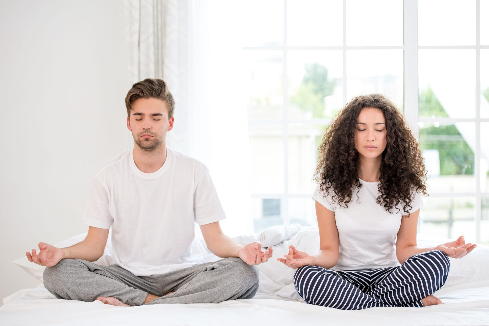 Mujer meditando sobre una esterilla en una habitación soleada con plantas y estantes llenos de frascos.