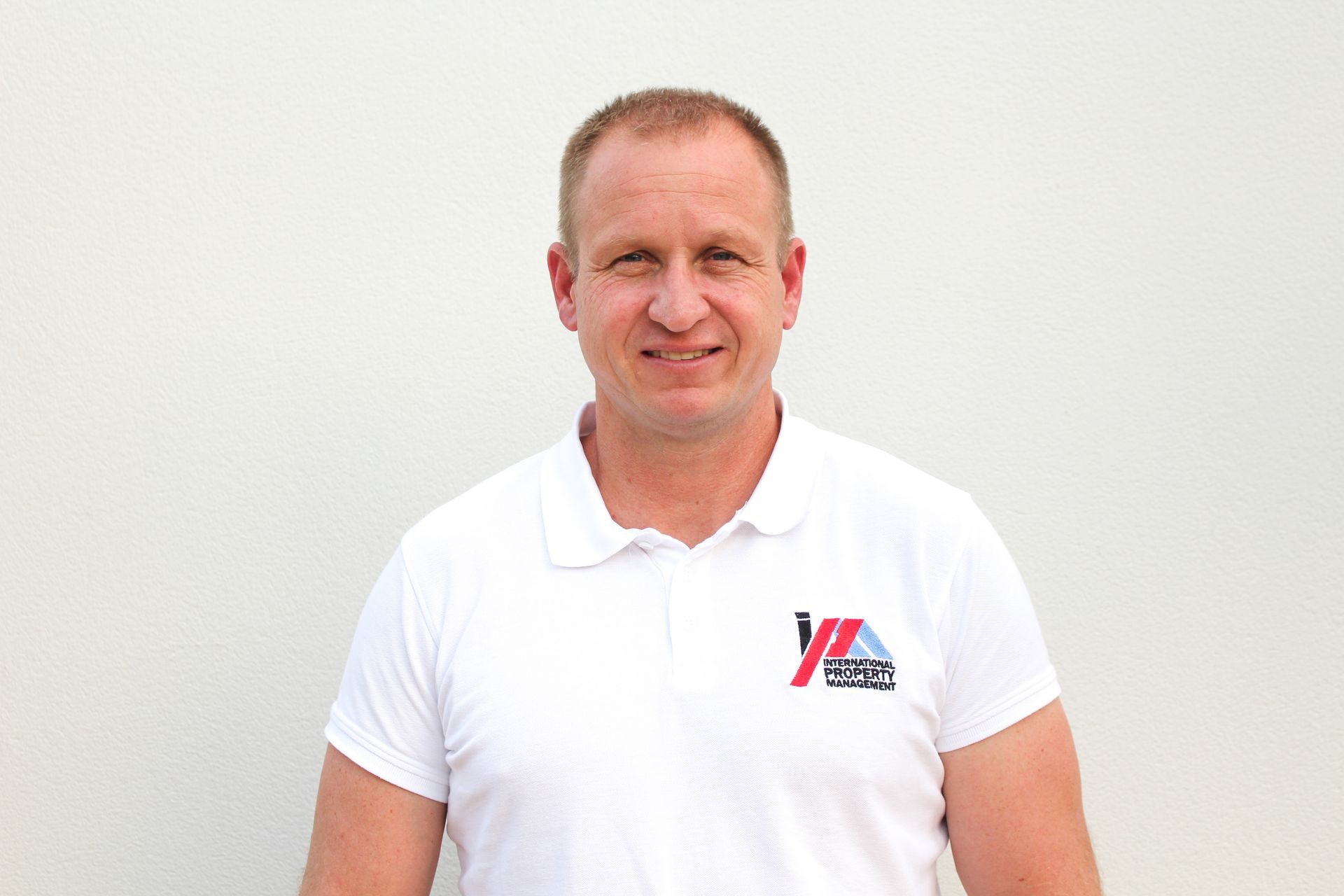 Un hombre vestido con un polo blanco y pantalones vaqueros está parado frente a una pared blanca.