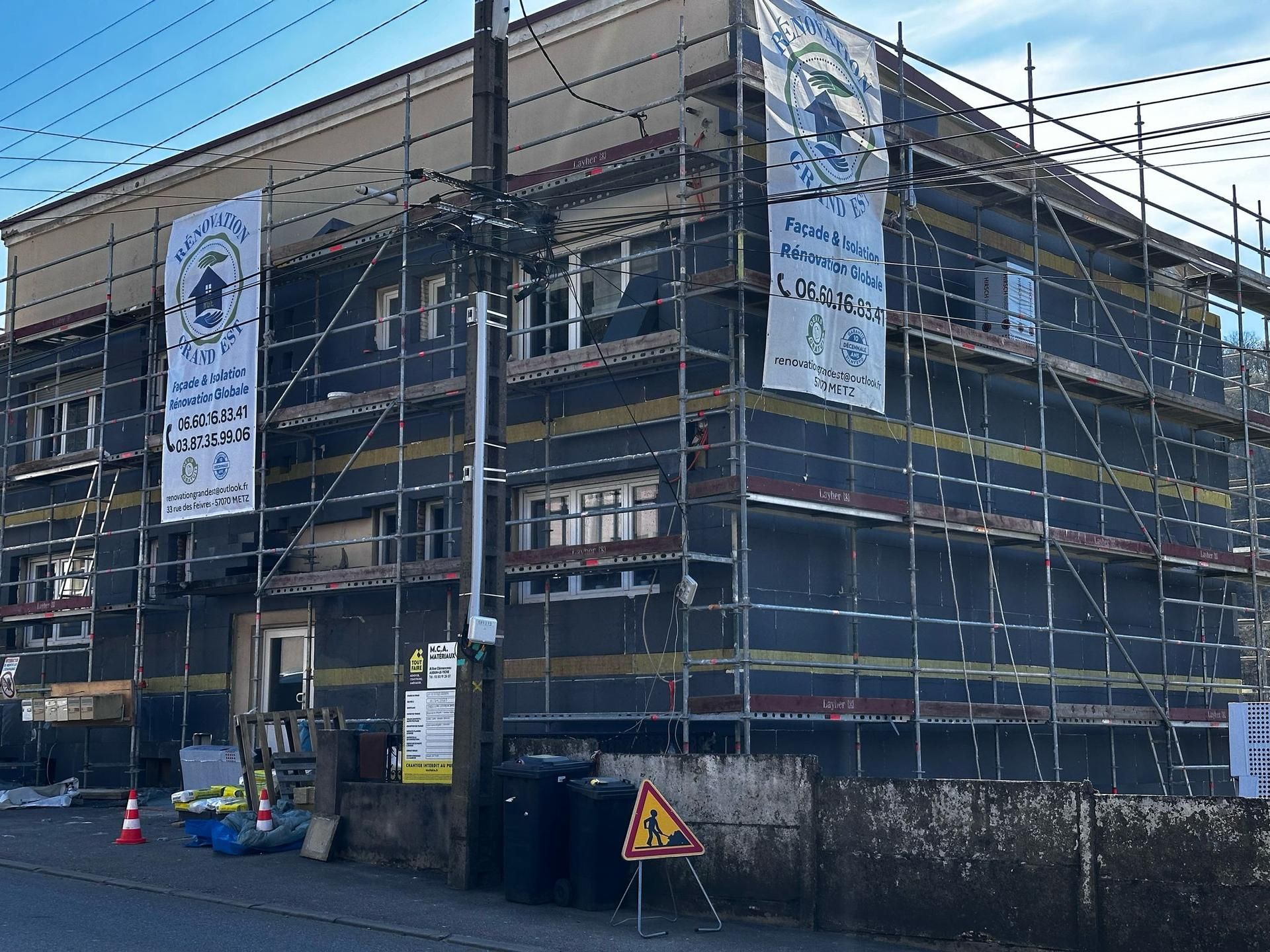 Bâtiment en rénovation avec échafaudages et isolation noire, banderoles et panneaux de signalisation.