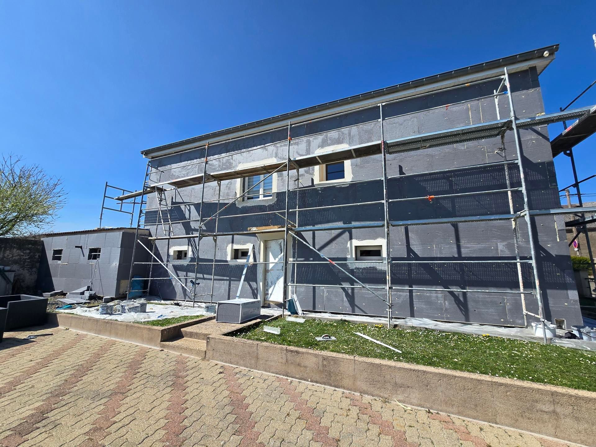 Extérieur d'un bâtiment en construction, échafaudages, panneaux isolants gris, ciel bleu.