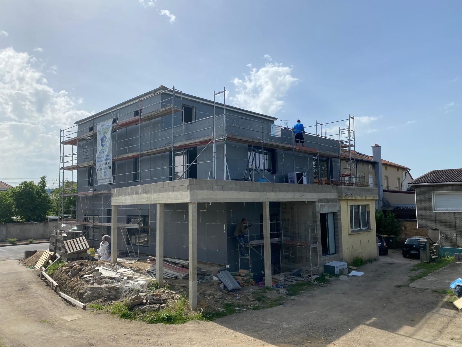 Maison à deux étages en construction avec échafaudage ; isolation grise, piliers en béton et un ouvrier sur le toit.