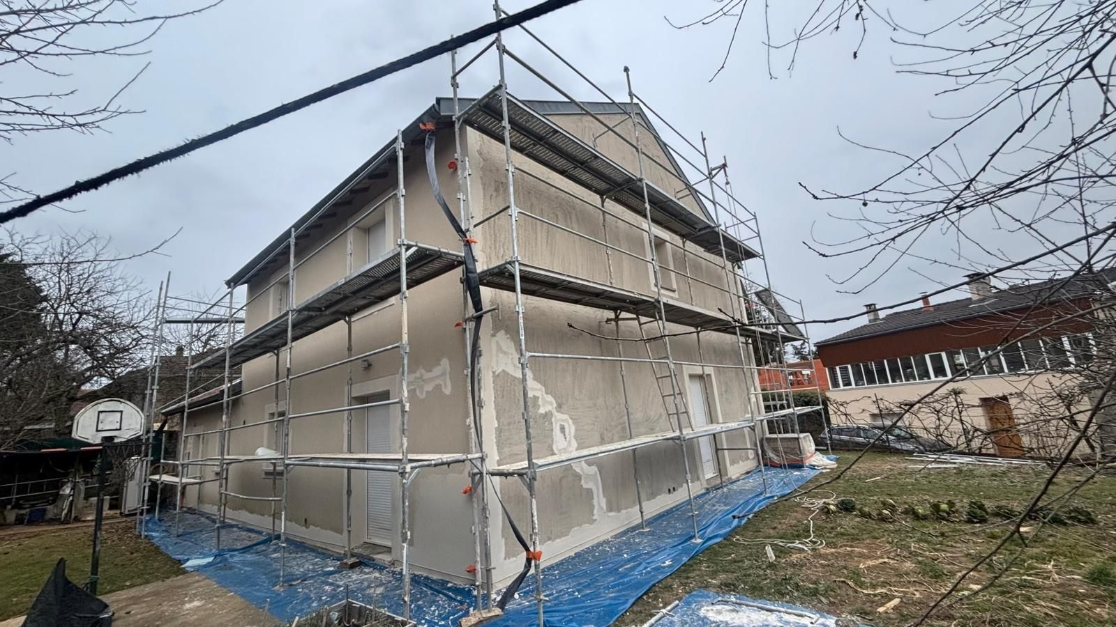 Extérieur de maison en rénovation avec échafaudage; bâche bleue, ciel couvert, arbres.
