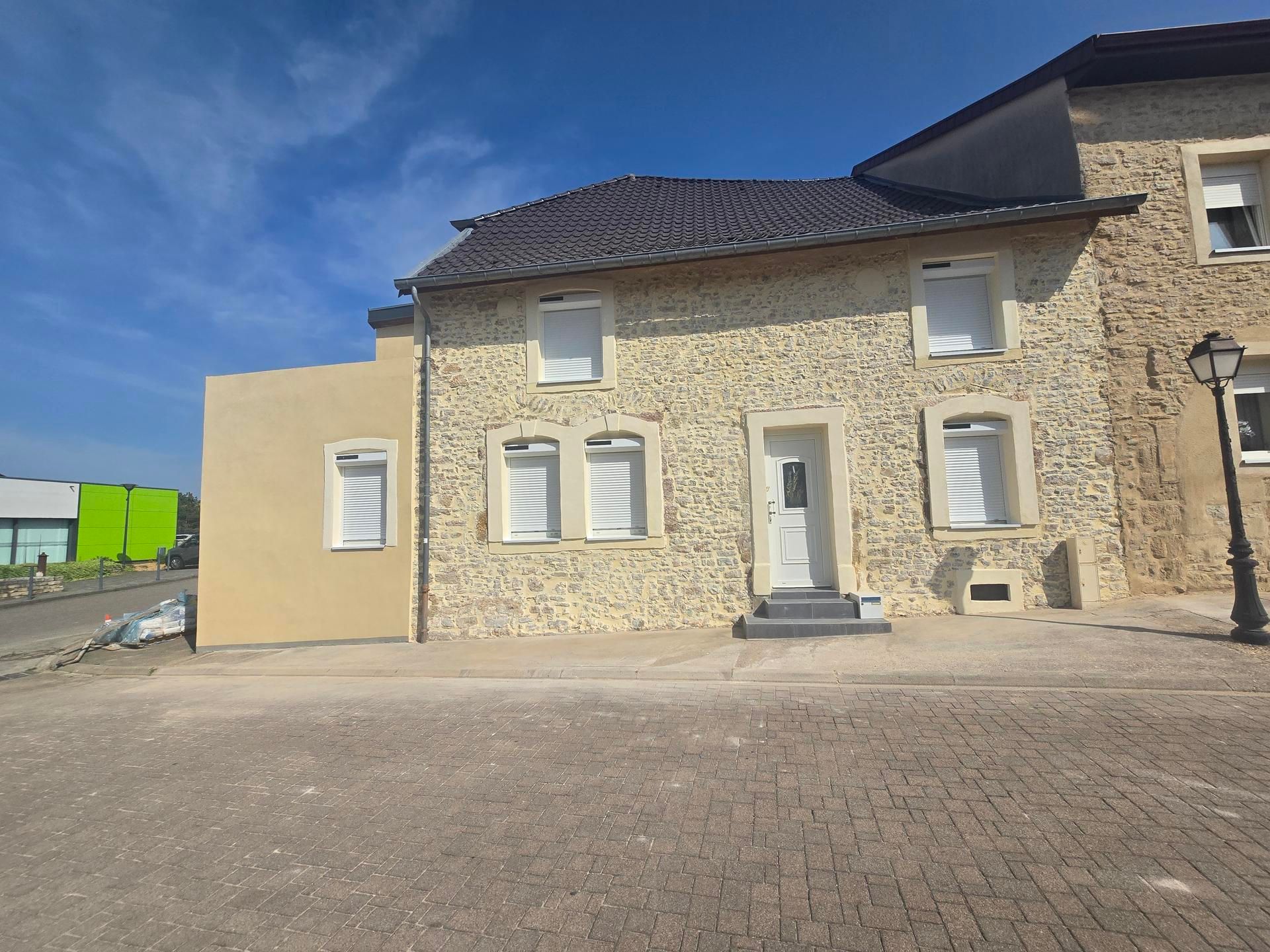 Maison à deux étages avec façade en stuc, volets blancs et toit en tuiles foncées.