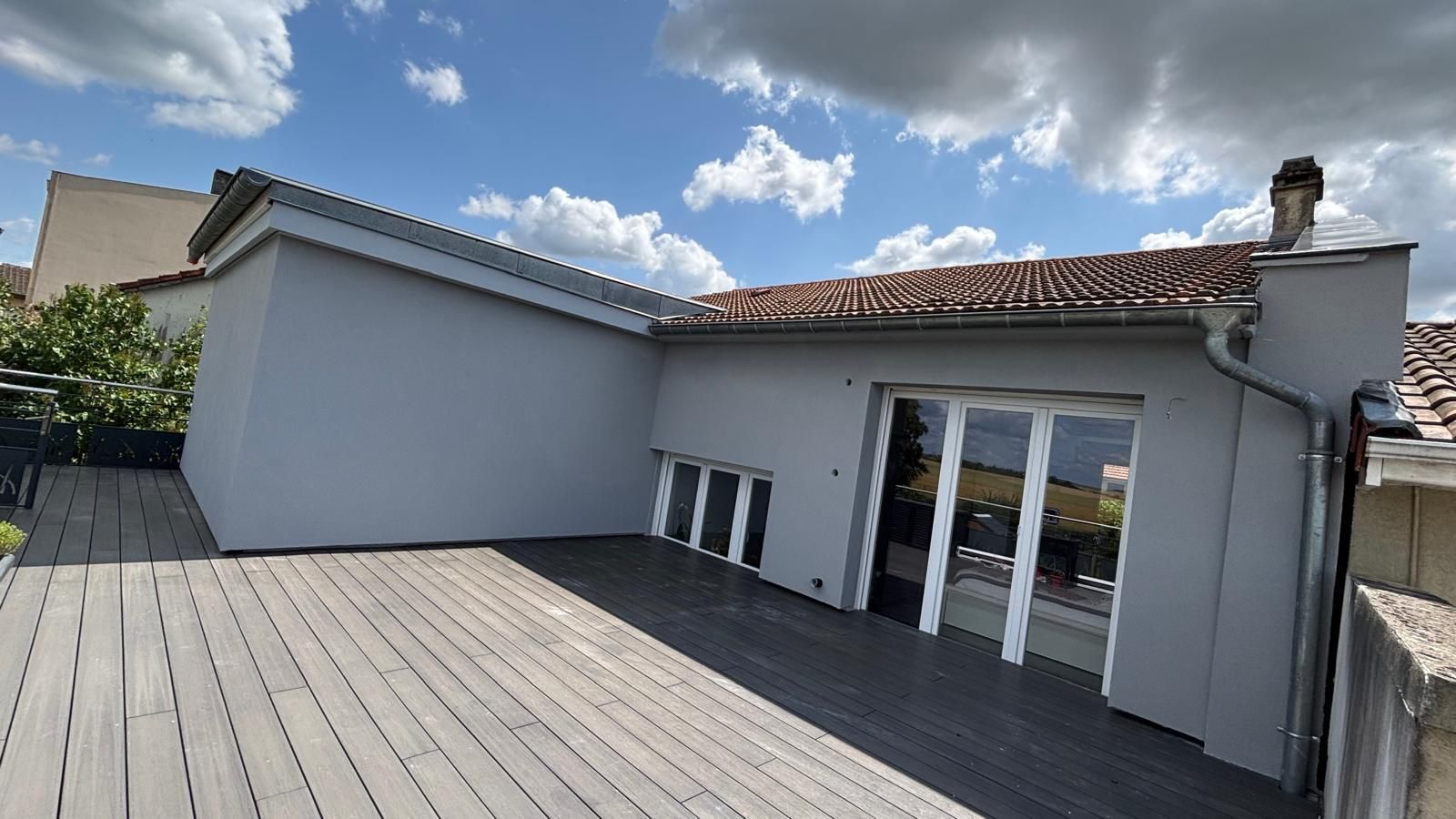 Un bâtiment gris avec une terrasse, des portes vitrées et un toit en tuiles par une journée ensoleillée.
