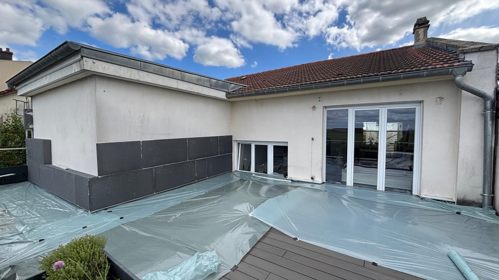 Vue extérieure d'une maison avec un toit plat recouvert de bâche en plastique, d'un revêtement gris et de portes vitrées.