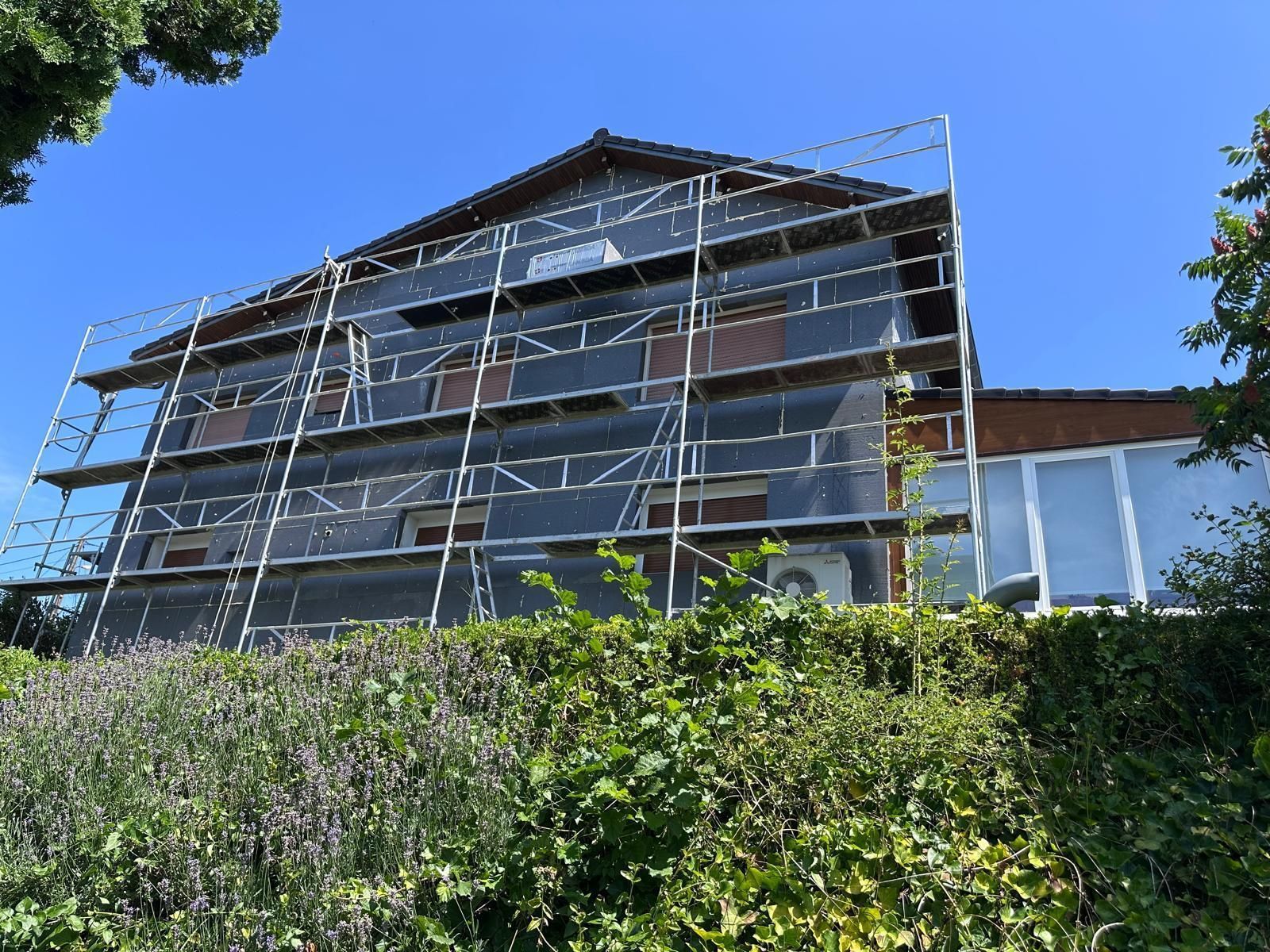 Maison avec échafaudage, recouverte de bâche noire, sous un ciel bleu, vue depuis un jardin.