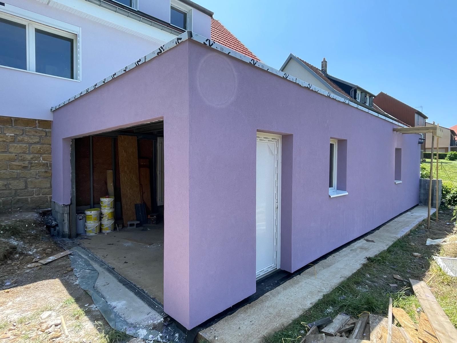 Extérieur d'un bâtiment nouvellement construit de couleur lavande avec un garage ouvert, une porte et des fenêtres sous un ciel bleu.