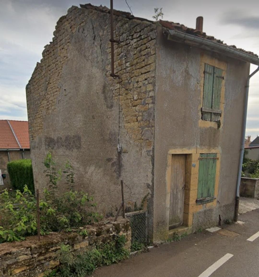 Un bâtiment de deux étages patiné avec des volets verts, une porte marron et des murs beiges en ruine à côté d'une route.