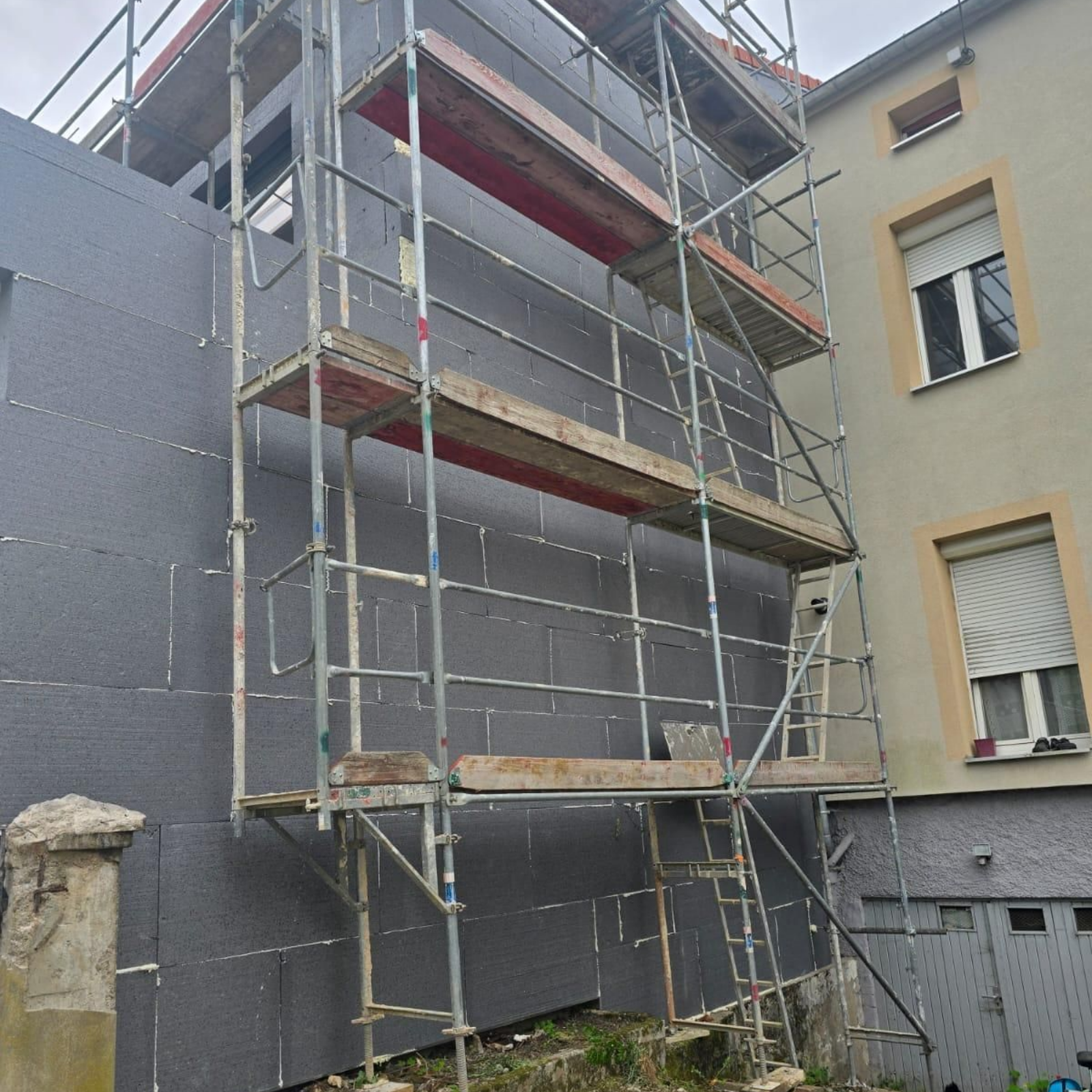 Échafaudage sur un bâtiment avec panneaux isolants foncés. Structures grises, rouges et en bois.