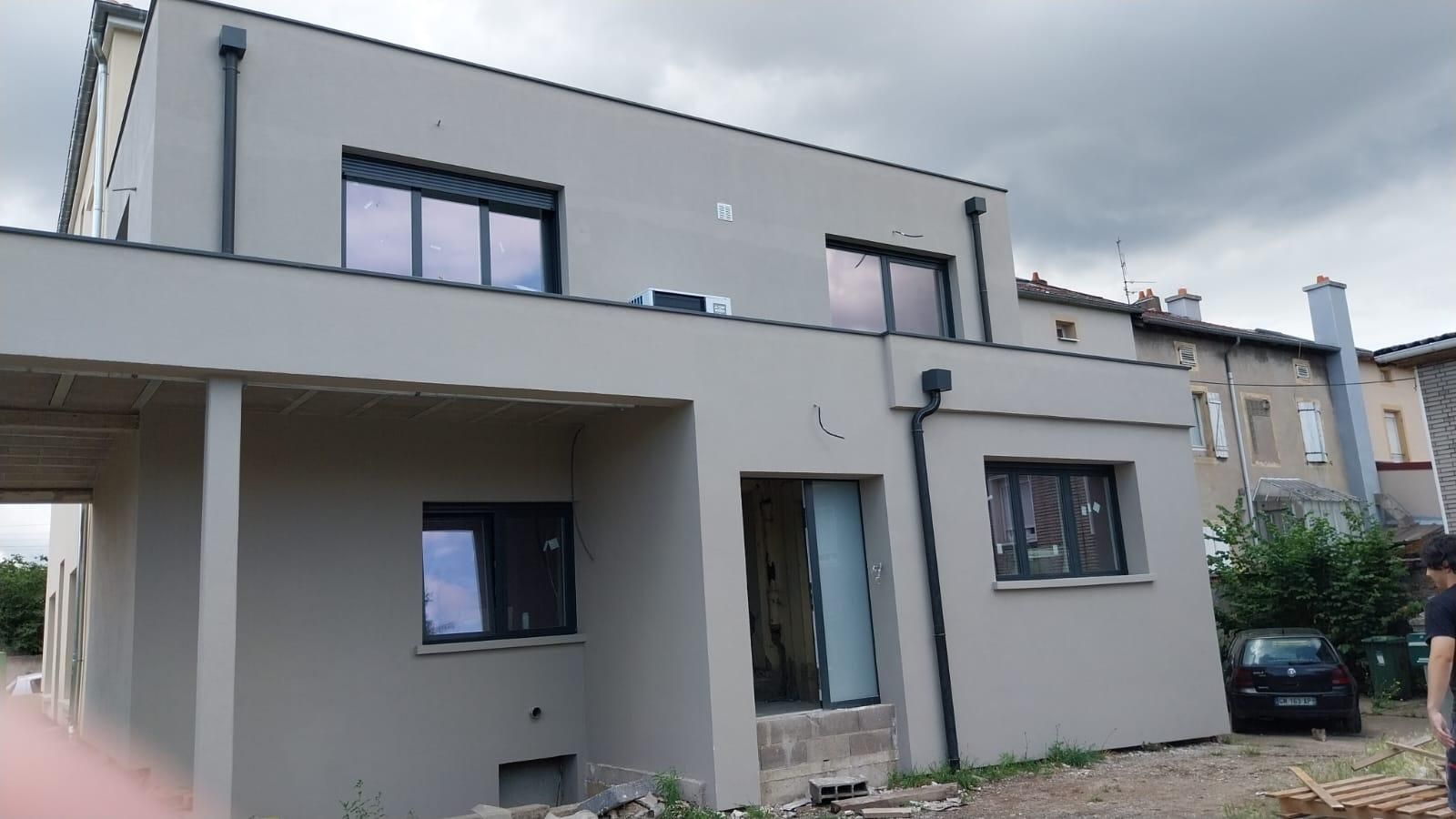 Maison moderne à deux étages avec extérieur gris, cadres de fenêtres noirs et ciel nuageux.