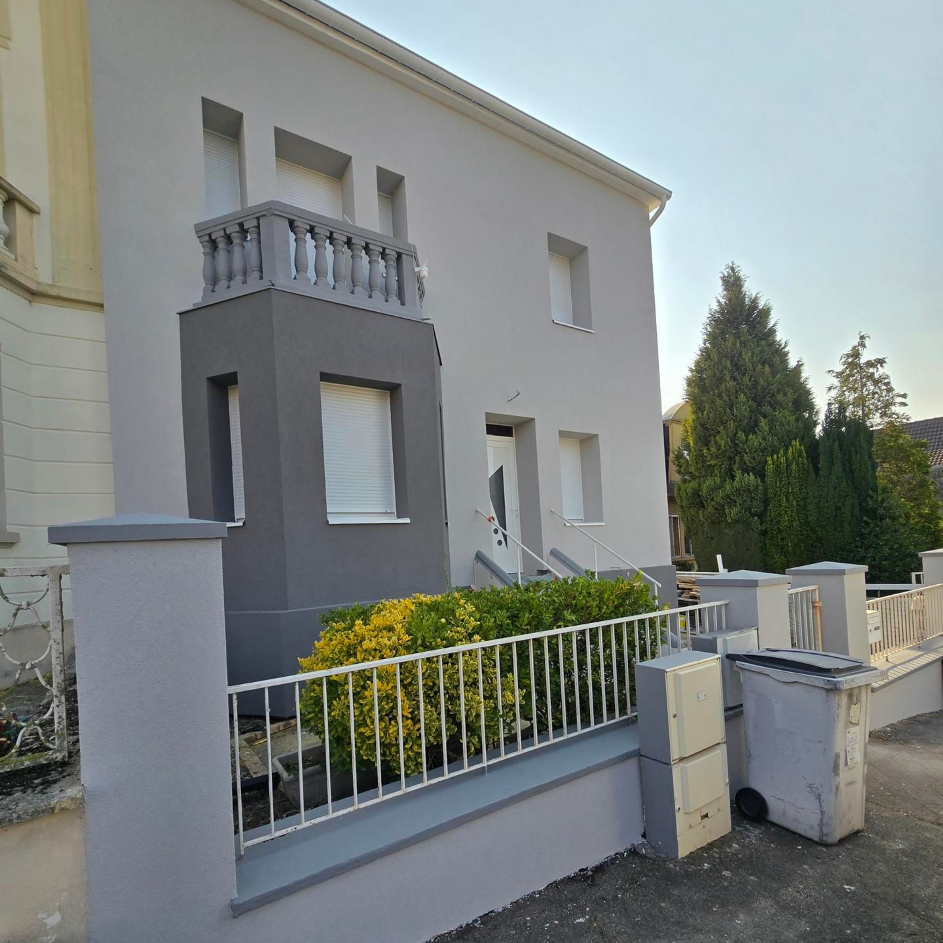 Maison grise à deux étages avec balcon, boiseries blanches et petite clôture.