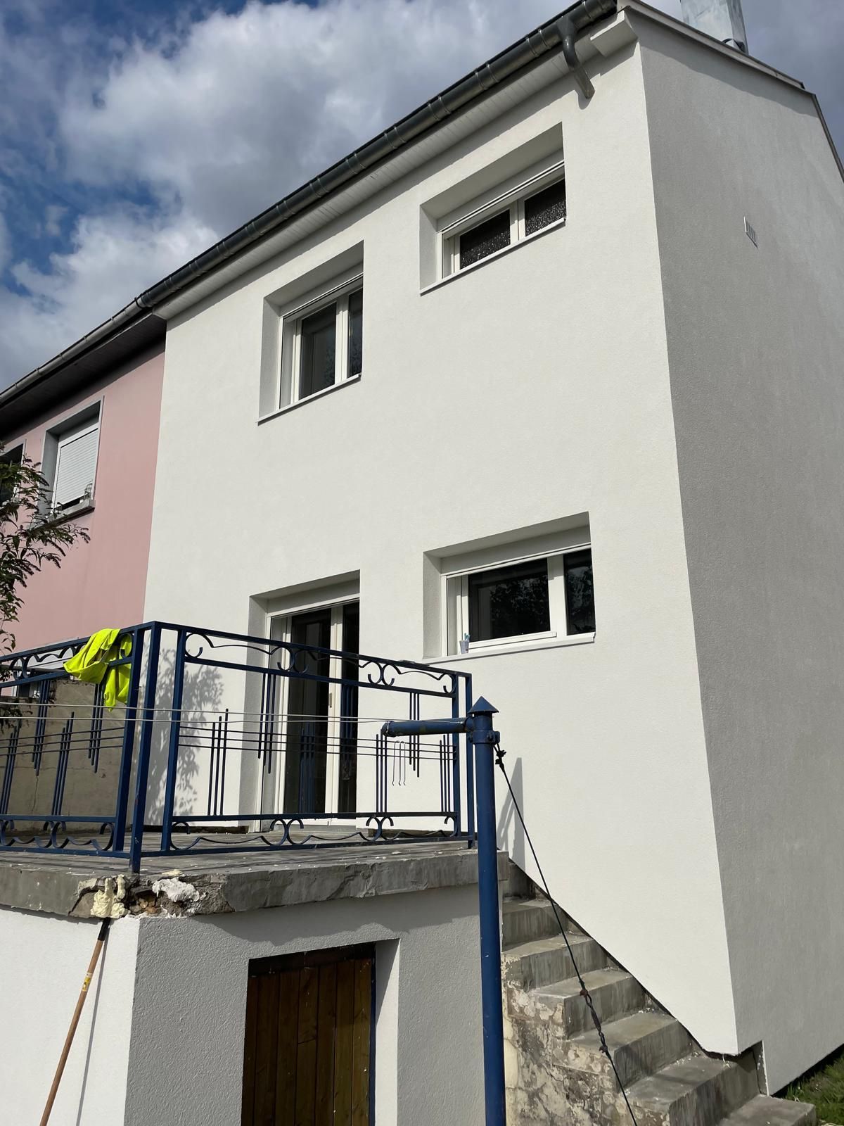 Maison blanche à deux étages avec fenêtres, balcon et escaliers, à côté d'un bâtiment rose.