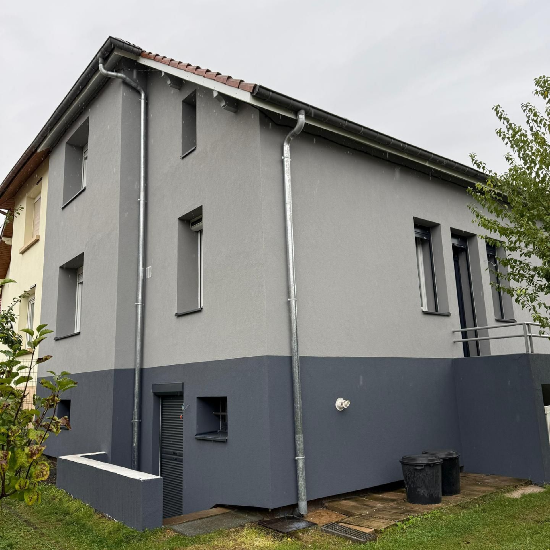 Maison à deux étages avec extérieur gris et gris foncé, gouttières et une petite cour par temps nuageux.