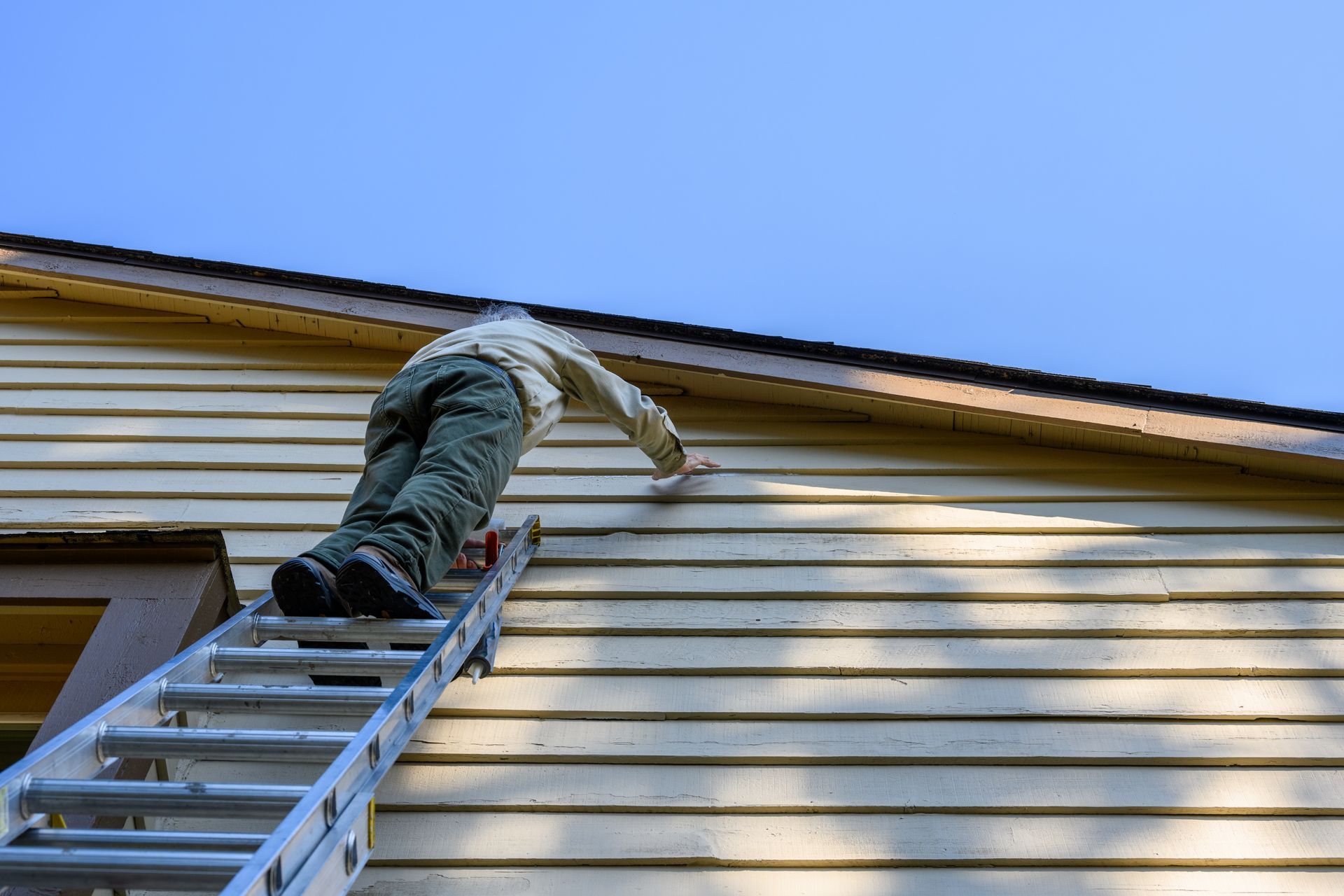 Une personne peint le revêtement d'une maison depuis une échelle, contre un ciel bleu.