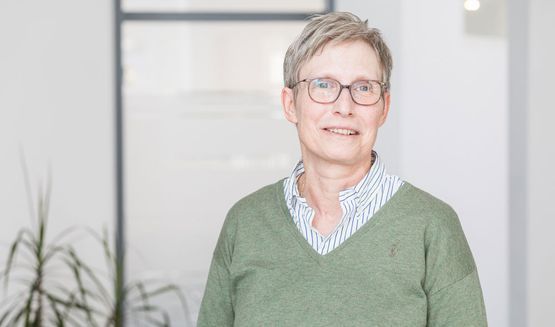 Eine Frau mit Brille und grünem Pullover steht vor einem Fenster.