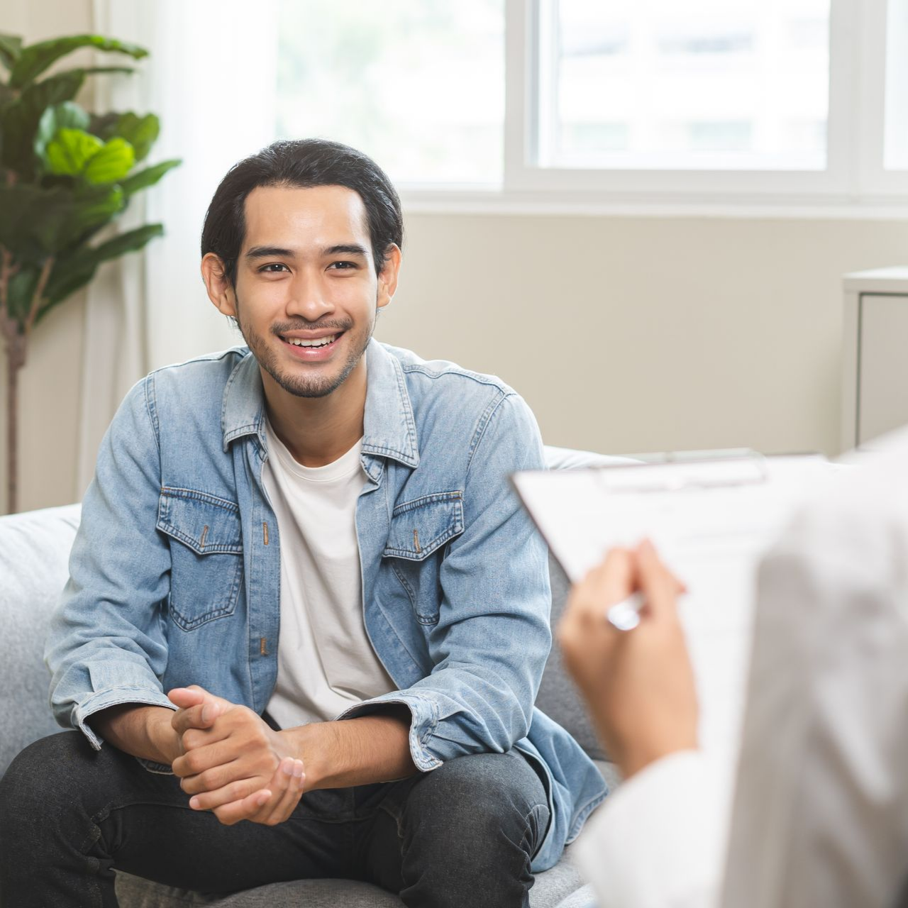 Un homme sourit pendant une consultation, assis sur un canapé devant une personne tenant un bloc-note.
