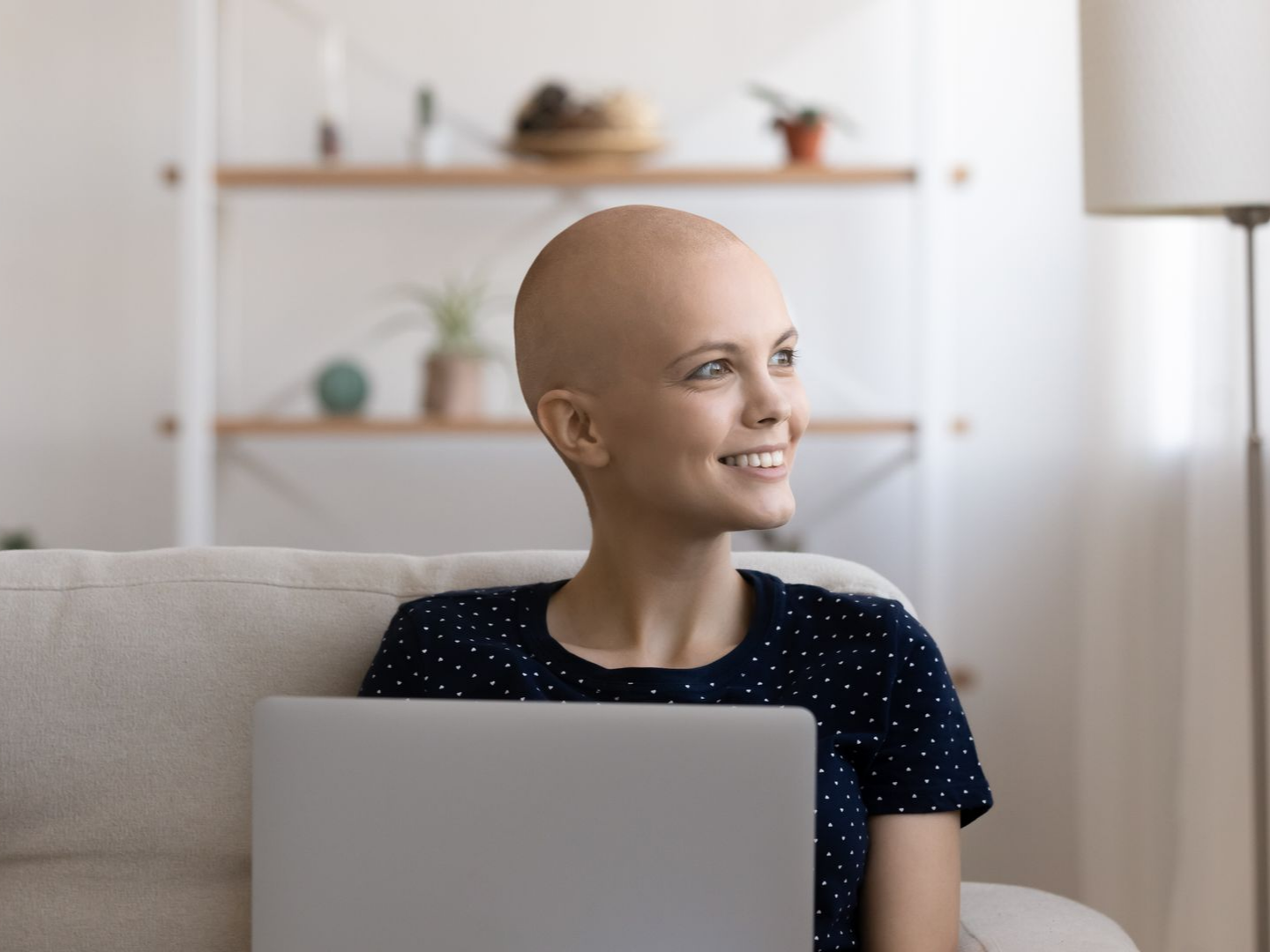 Une femme au crâne rasé sourit, tenant un ordinateur portable sur un canapé.