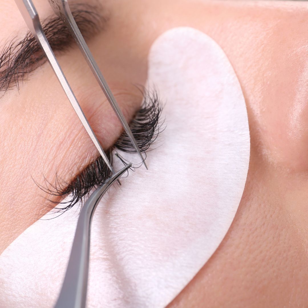 Extensiones de pestañas aplicadas al ojo de una persona con pinzas, sobre una almohadilla blanca.
