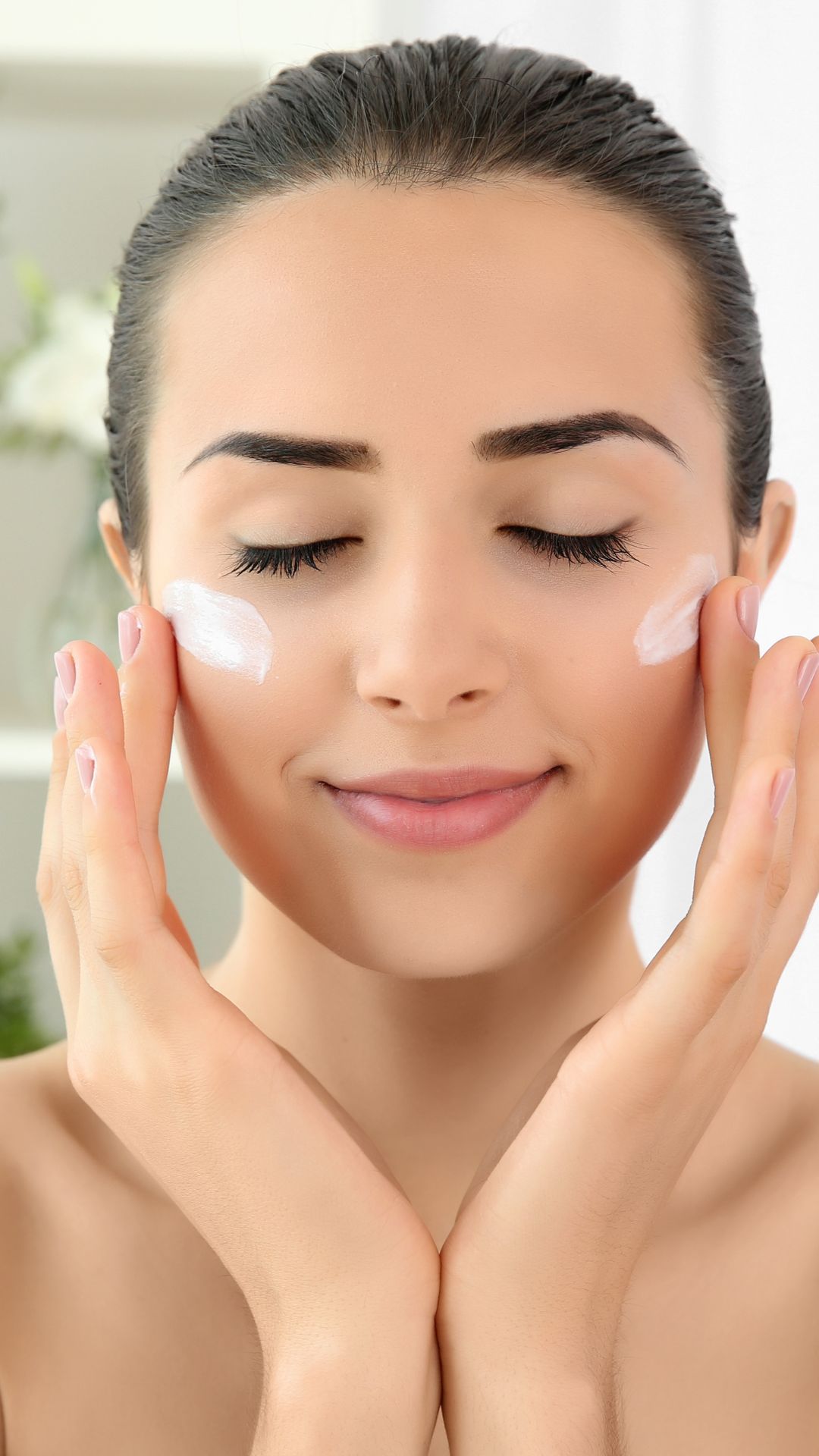 Mujer aplicándose crema hidratante en la cara con los ojos cerrados y las manos cerca de las mejillas.