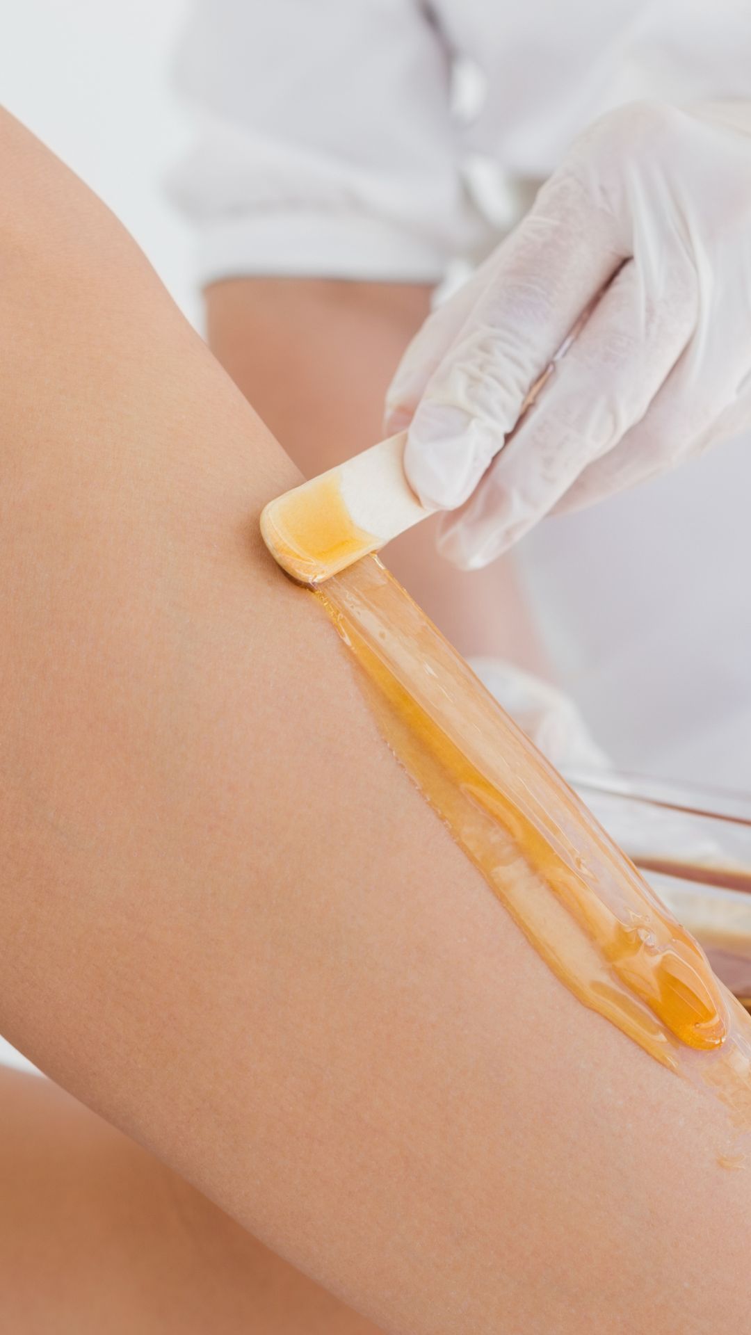 Una mano enguantada aplica cera tibia en una pierna con una espátula de madera, preparándola para la depilación.