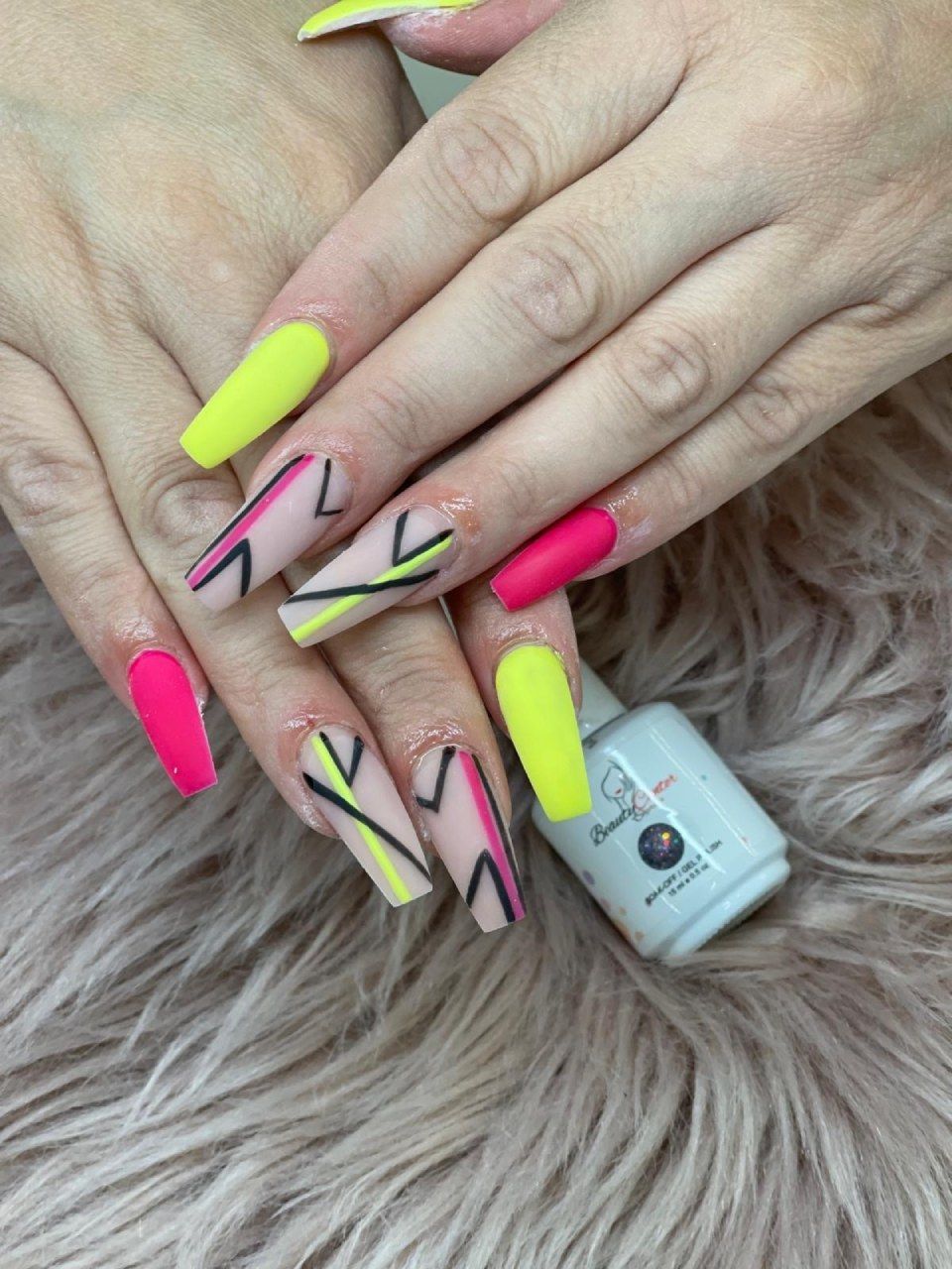 Hände mit leuchtend neonfarbenen Sargnägeln in rosa und gelben Mustern, die auf flauschigem Fell ruhen.