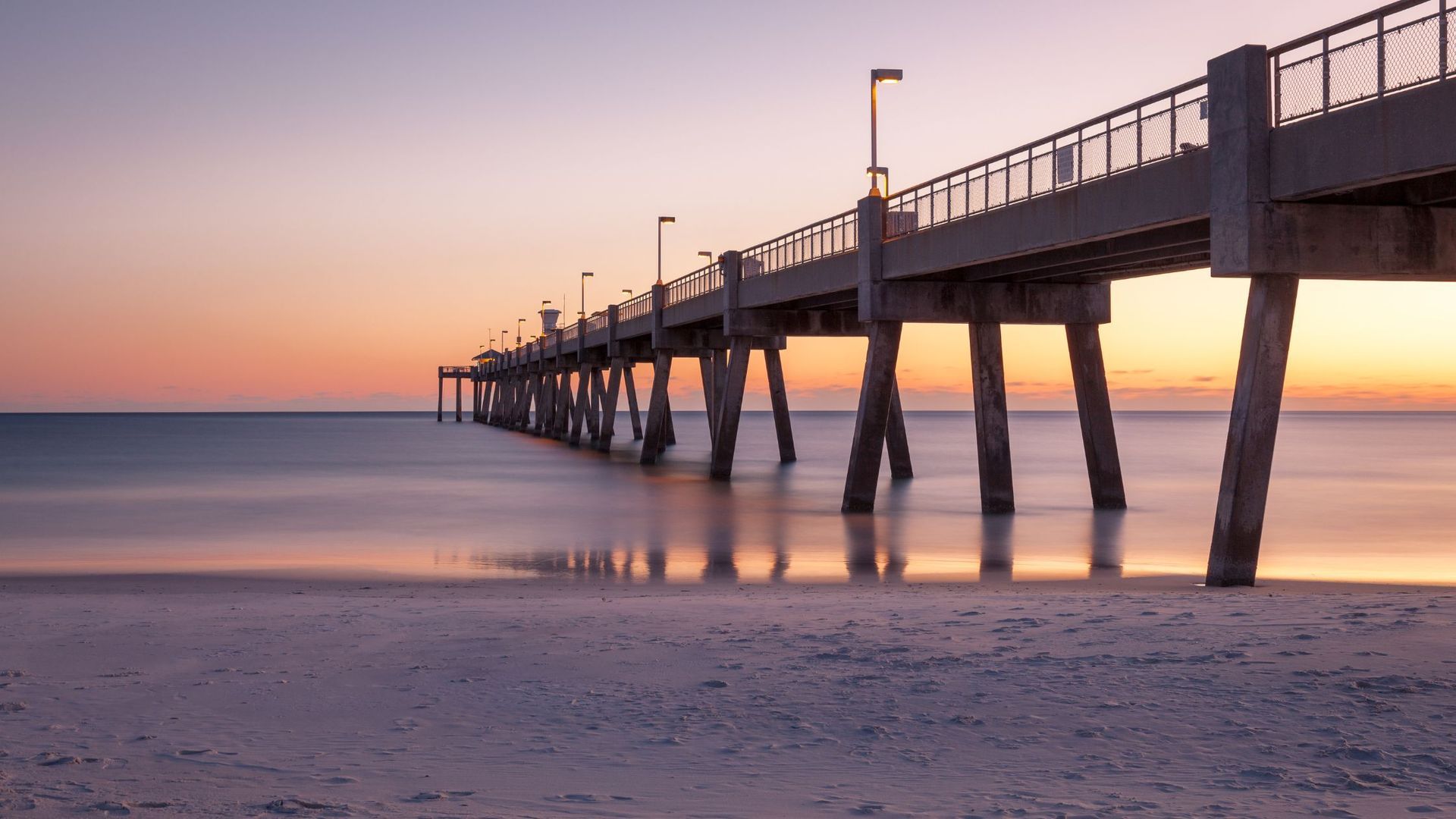 Pier extending over calm ocean at sunset; pink and orange sky reflecting on water and sand.