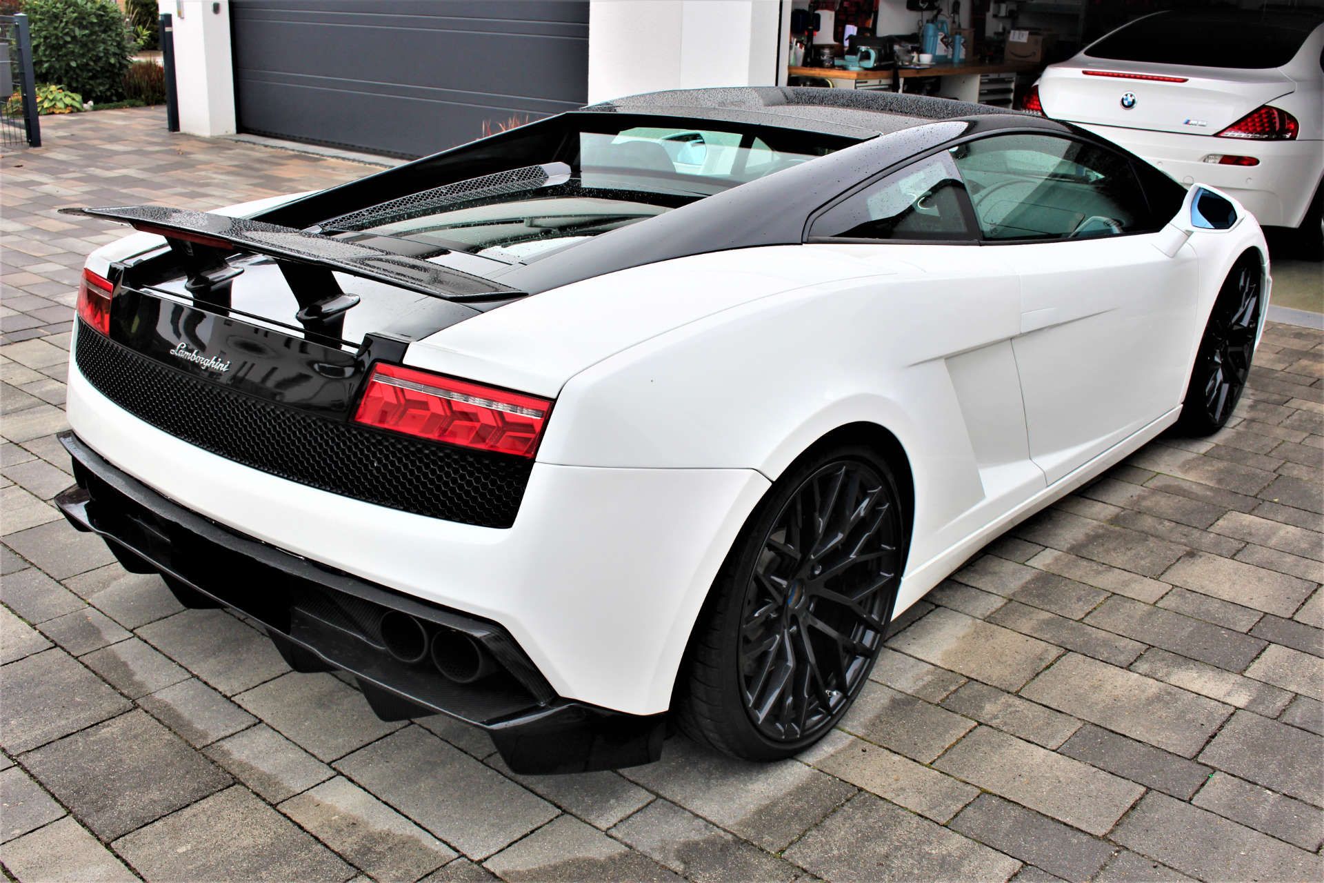 Weißer Lamborghini-Sportwagen mit schwarzem Dach und Heckspoiler, geparkt auf gepflasterter Einfahrt vor Garage.