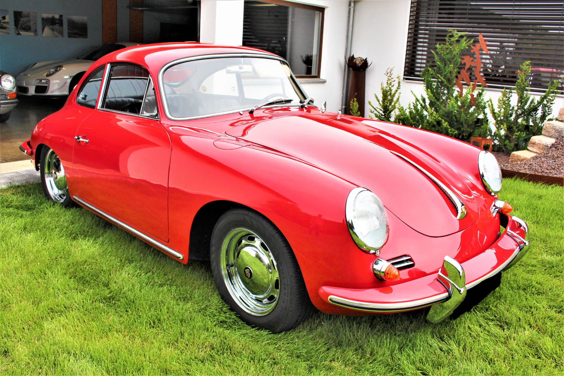 Roter Porsche 356 Coupé mit Chromfelgen auf grünem Rasen vor einem Haus mit Fenster und Pflanzen.