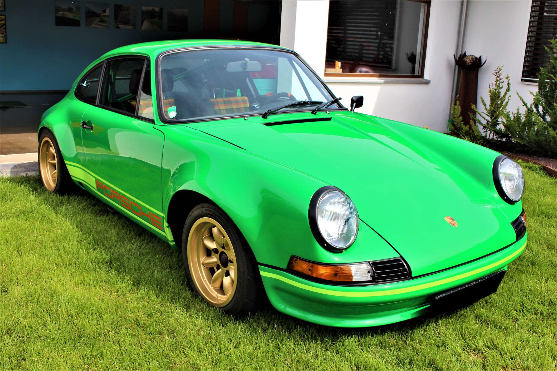 Grüner Porsche 911 mit goldfarbenen Felgen steht auf Rasen vor einem Haus mit weißen Wänden und Fenstern.