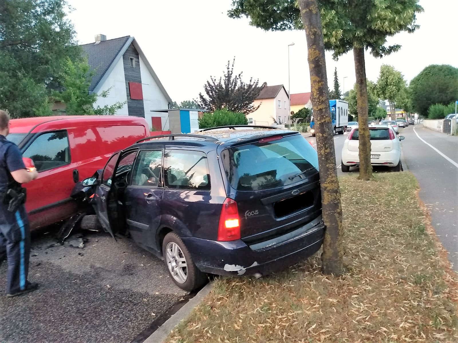 Blauer Ford Focus Kombi mit beschädigter Front und Heck steht neben einem Baum am Straßenrand, roter Transporter daneben.