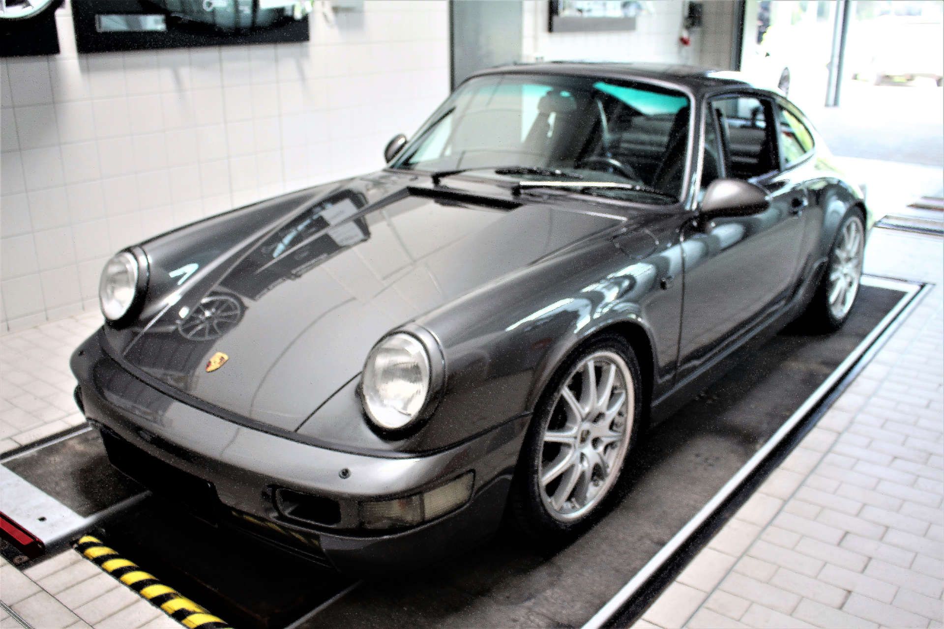 Dunkelgrauer Porsche 911 mit runden Scheinwerfern und silbernen Felgen in einer Werkstatt.
