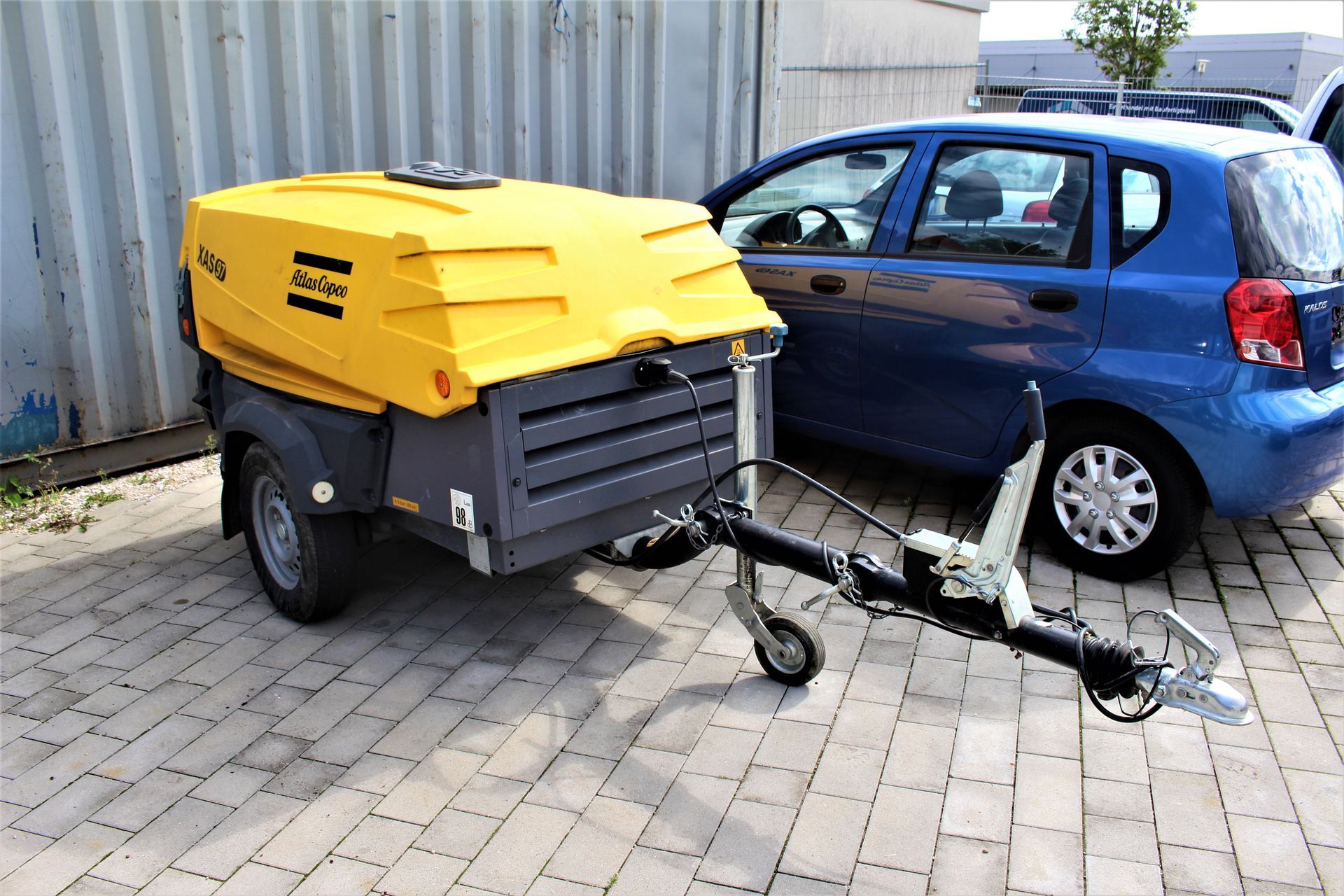 Gelb-schwarzer Anhängerkompressor der Marke Atlas Copco neben blauem Kleinwagen auf gepflastertem Hof.