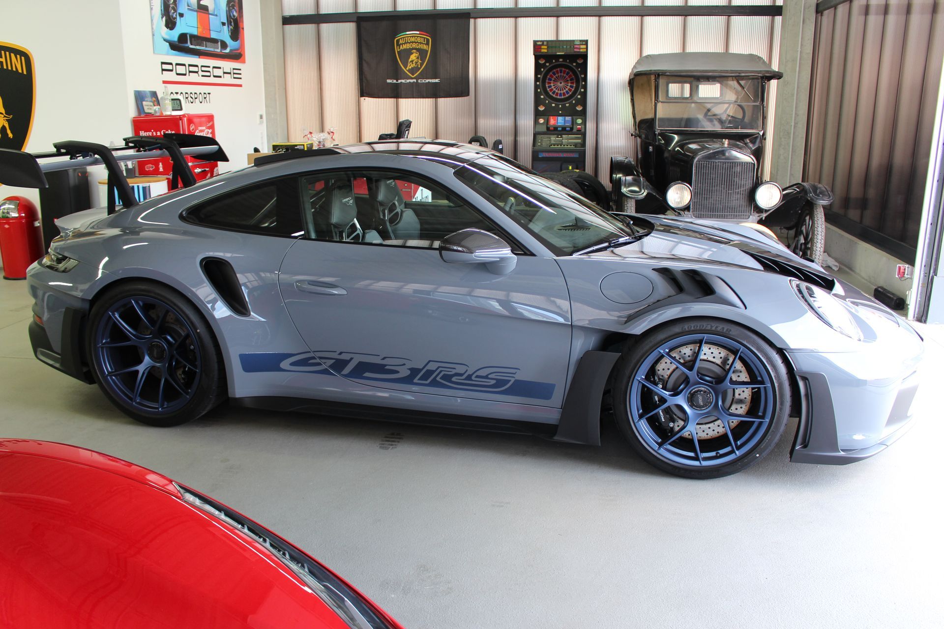 Seitliche Ansicht eines silbernen Porsche 911 GT3 RS mit blauen Felgen in einer Garage mit Oldtimer und Automarken-Postern.