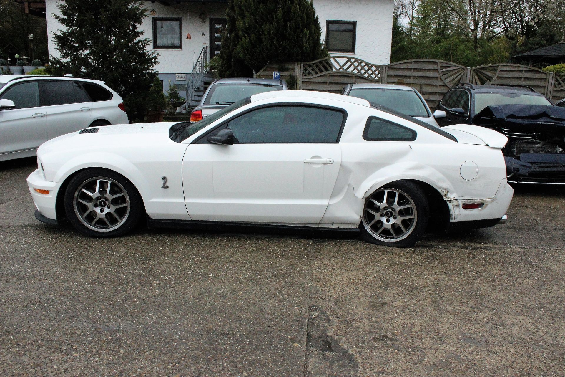 Weißer Ford Mustang mit Cobra-Emblem, beschädigtem hinterem Kotflügel und abgeflachtem Hinterreifen, geparkt vor Wohnhäusern.