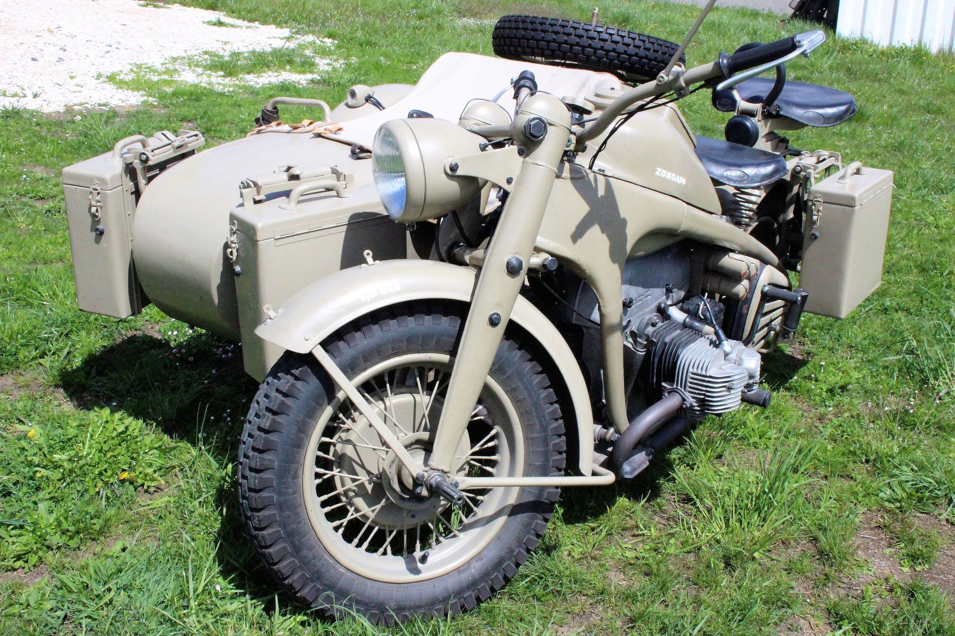 Beige Motorrad mit Beiwagen, zwei rechteckigen Koffern und einem Ersatzreifen auf dem Beiwagen, steht auf Gras.