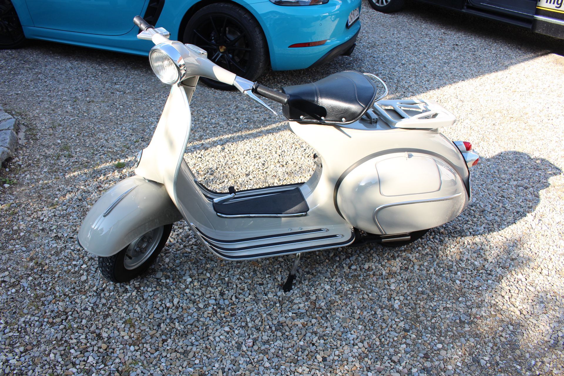 Beigefarbener klassischer Motorroller mit schwarzem Sitz auf kiesigem Untergrund, im Hintergrund blaue und schwarze Autos.