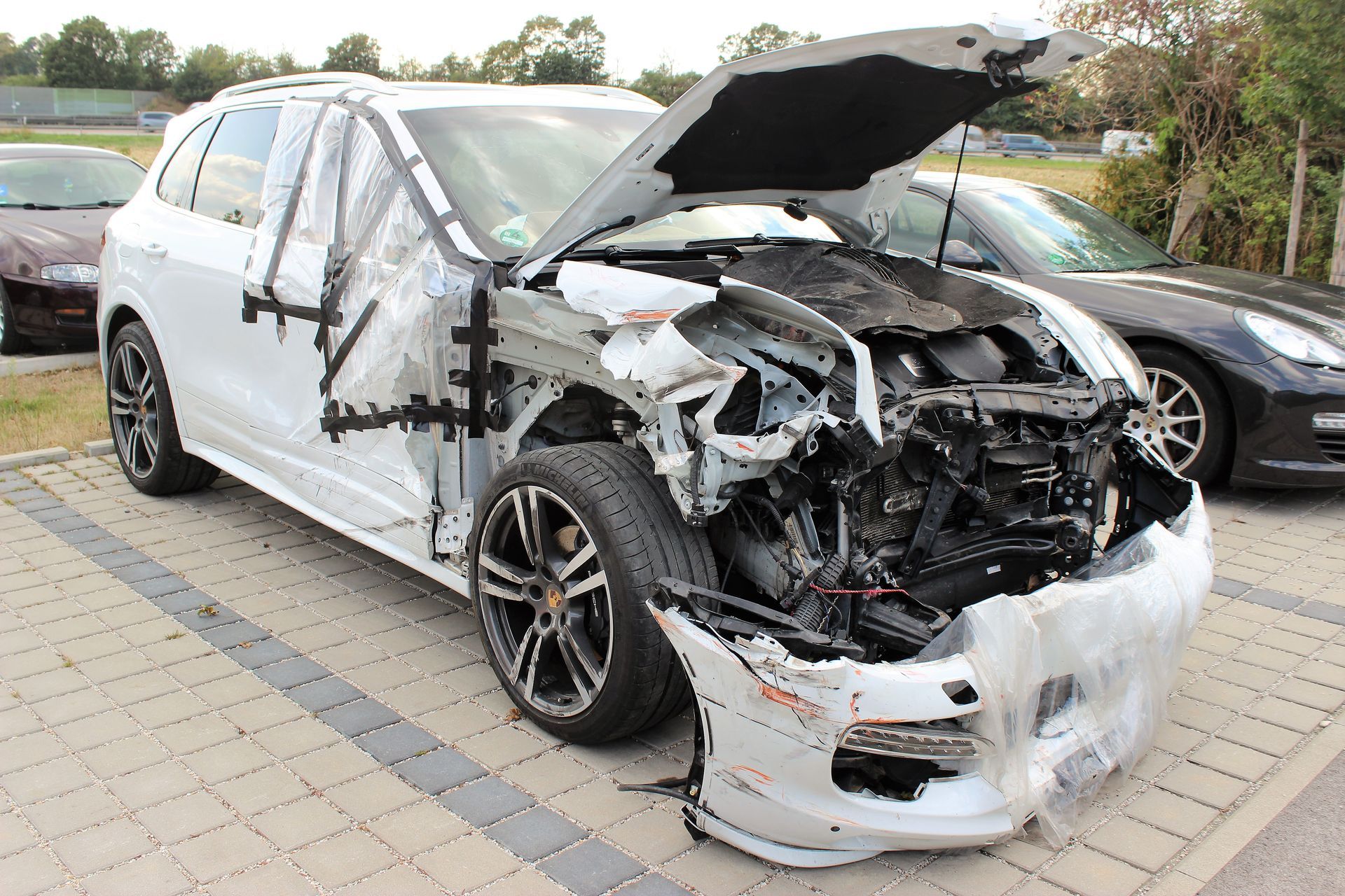 Weißer SUV mit stark beschädigter Front und rechter Seite, Motorhaube geöffnet, auf gepflastertem Parkplatz.