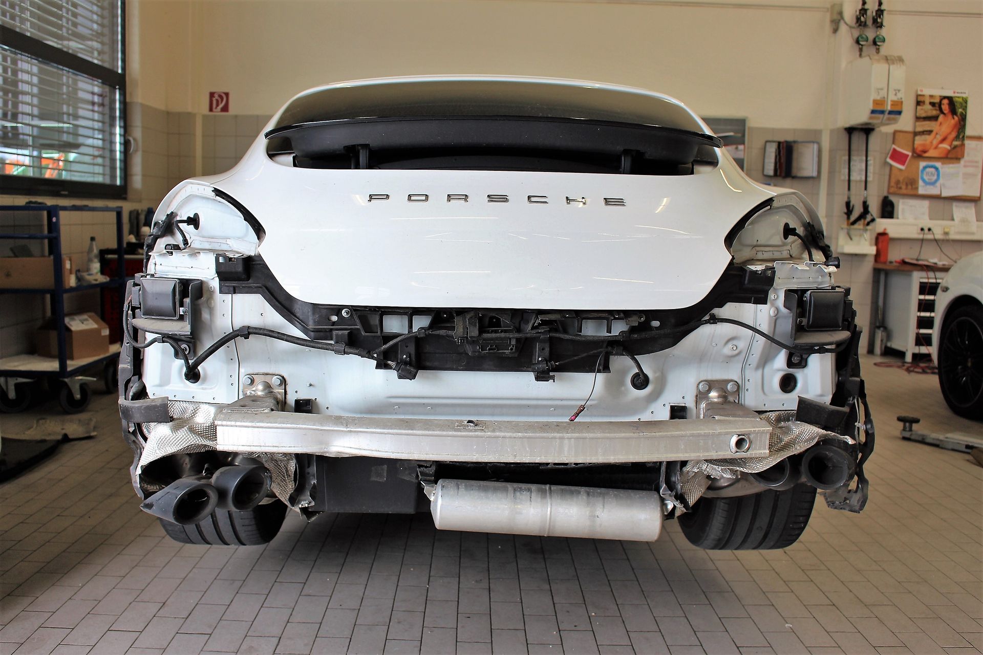 Heckansicht eines weißen Porsche ohne Stoßstange in einer Werkstatt mit sichtbarem Auspuff und freiliegenden Kabeln.