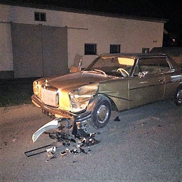 Goldfarbener Mercedes-Benz mit stark beschädigter Front und abgerissenem Stoßfänger auf einer Straße bei Nacht.