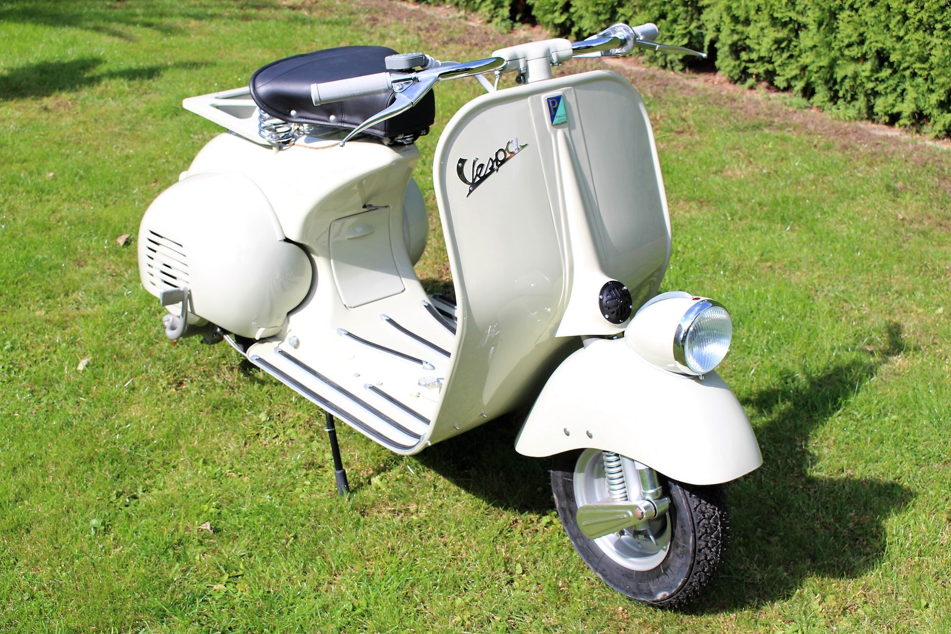 Beige Vespa-Roller mit schwarzem Sitz auf grünem Rasen, Seitenständer ausgeklappt.
