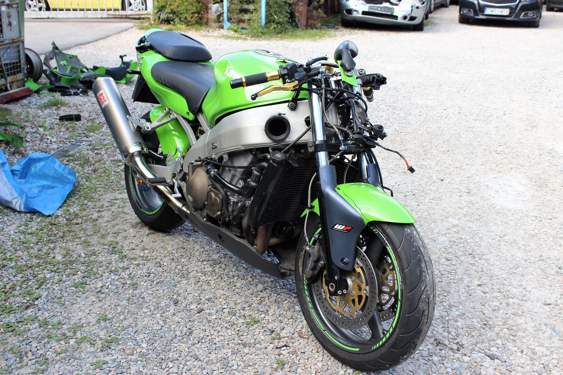 Grünes Motorrad ohne Verkleidung, sichtbar Motor und Rahmen, steht auf Kiesboden, im Hintergrund Autos und Werkstattbereich.