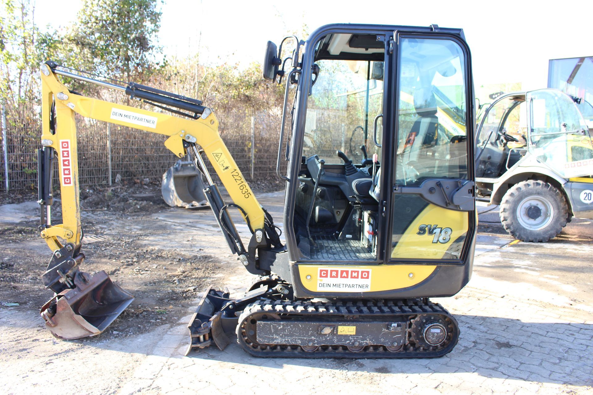 Gelber Minibagger mit Kettenfahrwerk und geschlossener Fahrerkabine steht auf gepflastertem Untergrund.