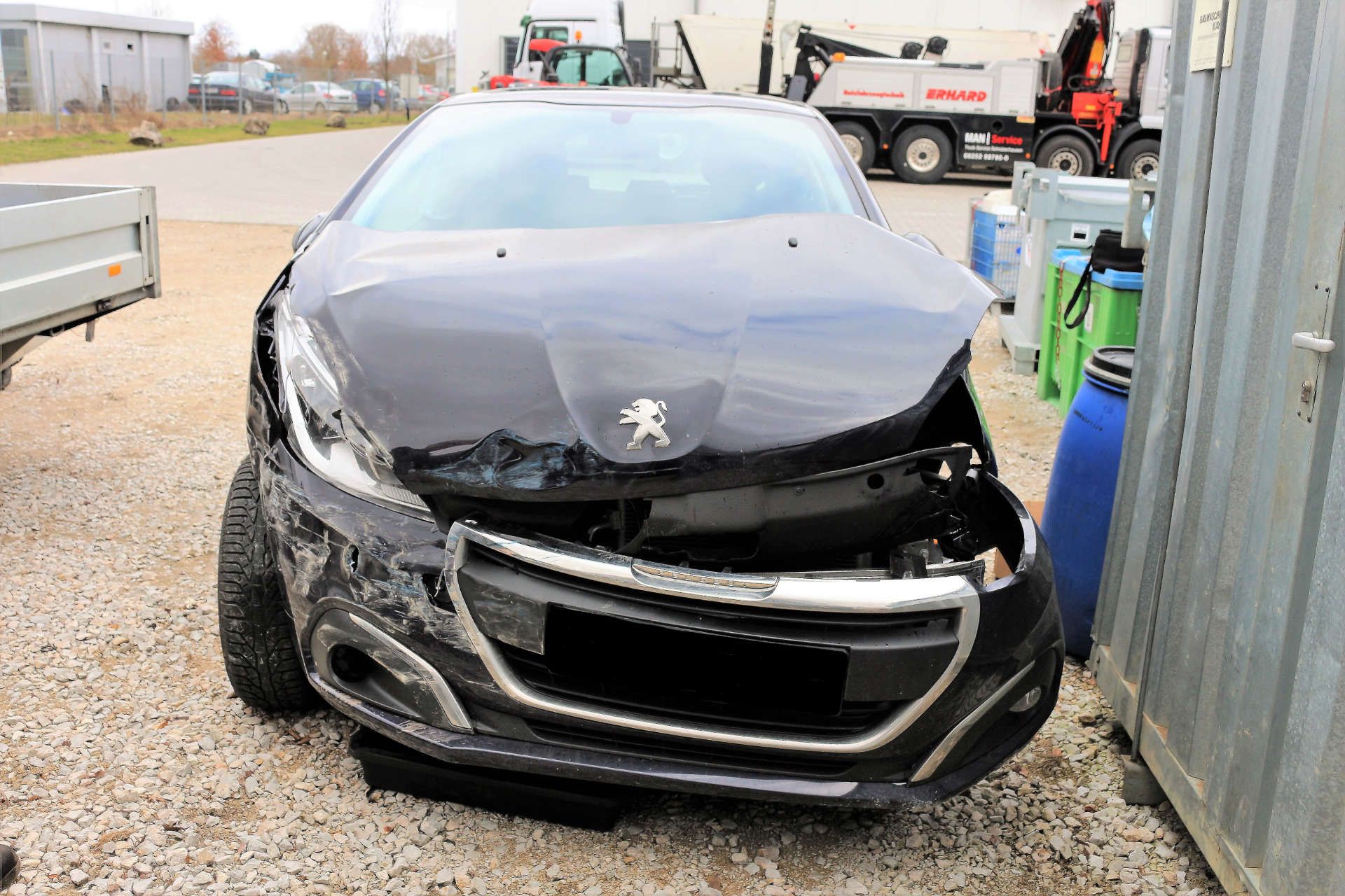 Peugeot-Auto mit stark beschädigter Frontpartie, verbeulter Motorhaube und verschobener Stoßstange, geparkt auf Schotter.
