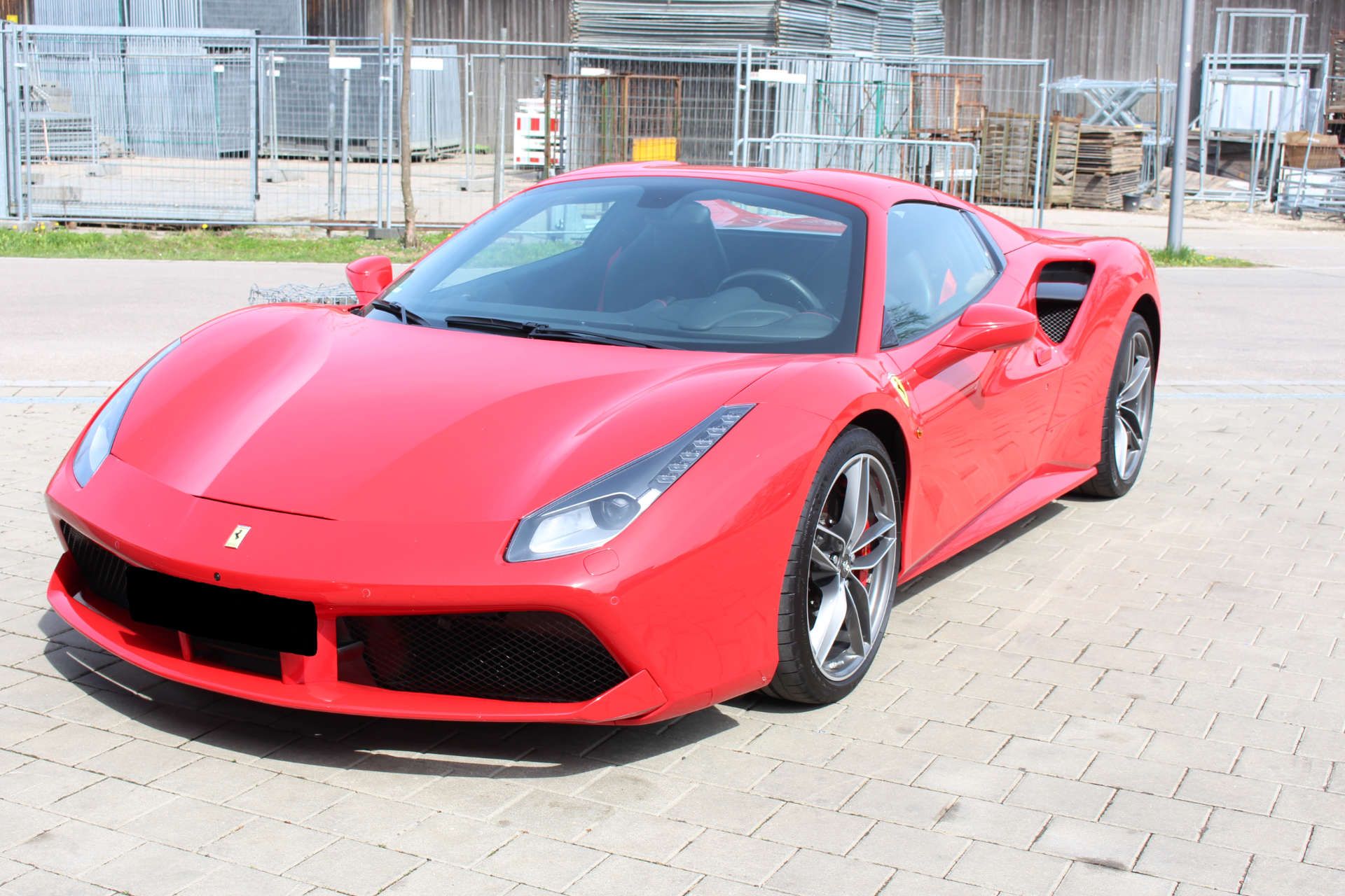 Roter Ferrari-Sportwagen mit deutschem Kennzeichen, geparkt auf gepflastertem Platz vor Baustellenabsperrung.
