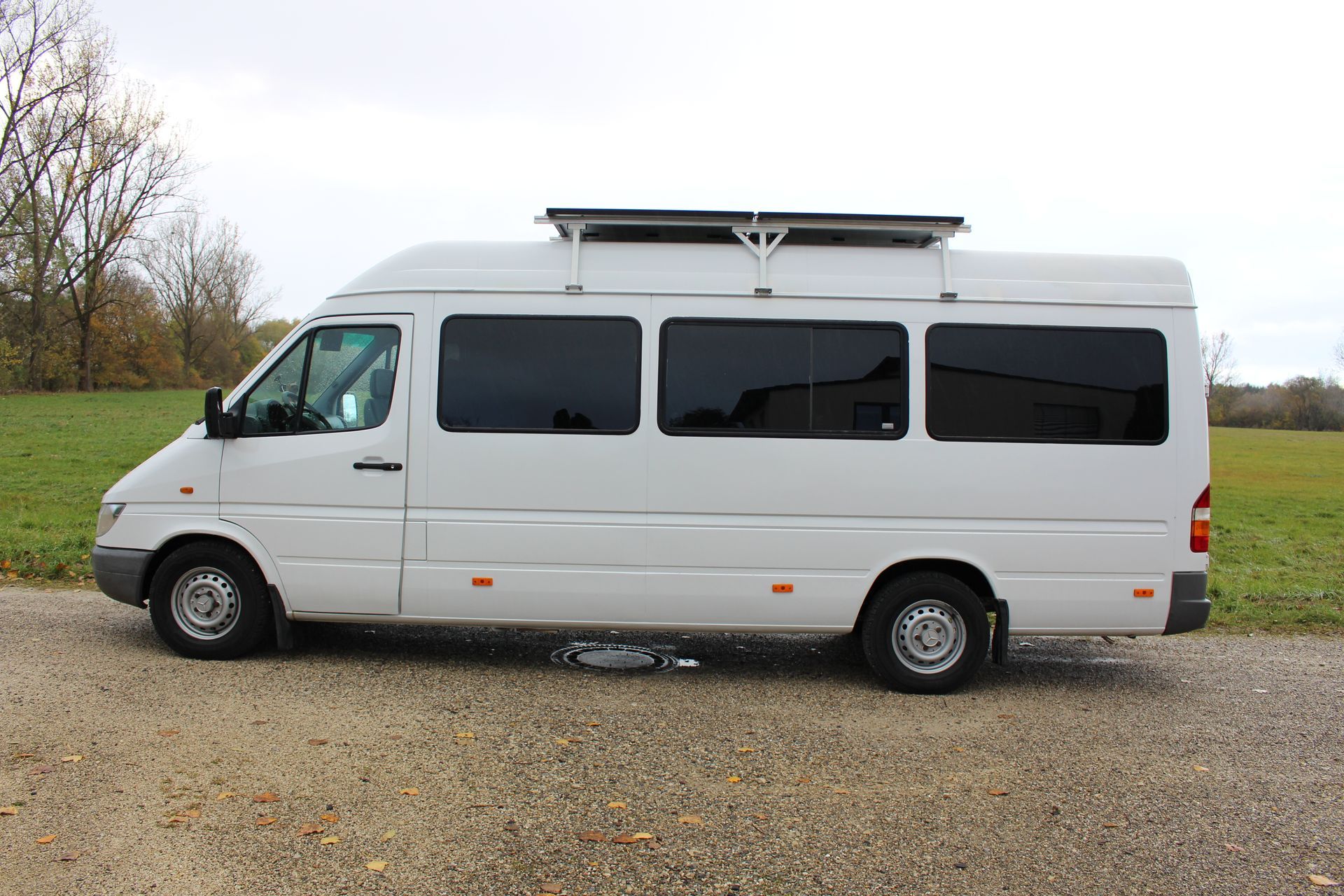 Weißer Transporter mit vier getönten Fenstern und Solarpanel auf dem Dach, geparkt auf Asphalt vor Wiese.