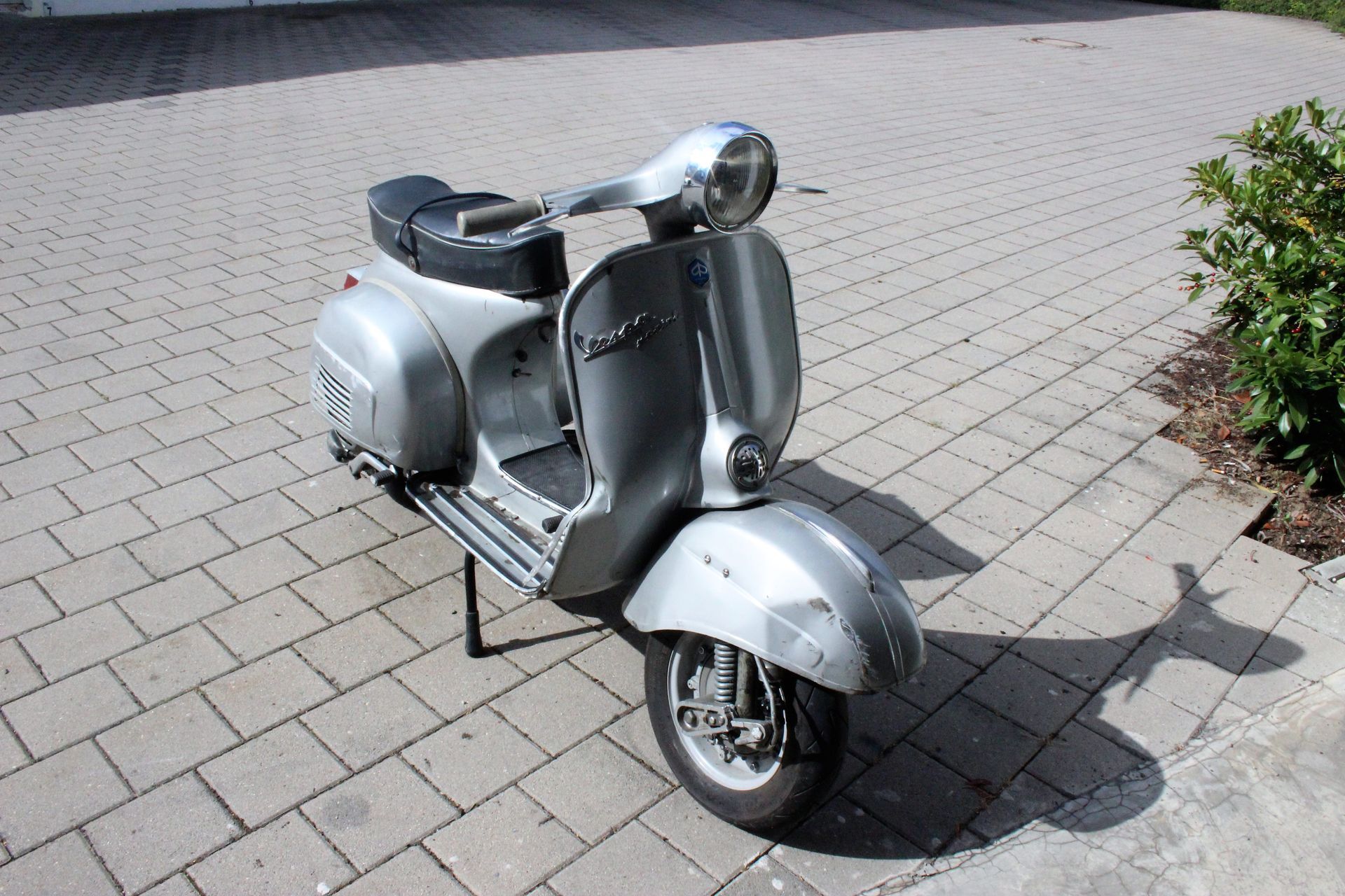 Silberner Vespa-Roller mit schwarzem Sitz auf gepflastertem Boden, im Sonnenlicht stehend.
