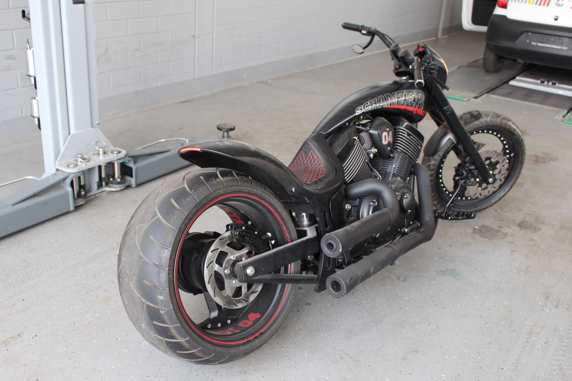 Schwarzes Motorrad mit breitem Hinterreifen, rotem Zierstreifen und auffälligem Auspuff in einer Garage.