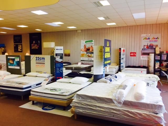 Salle d'exposition de matelas présentant plusieurs lits. Murs beiges, éclairage lumineux au plafond